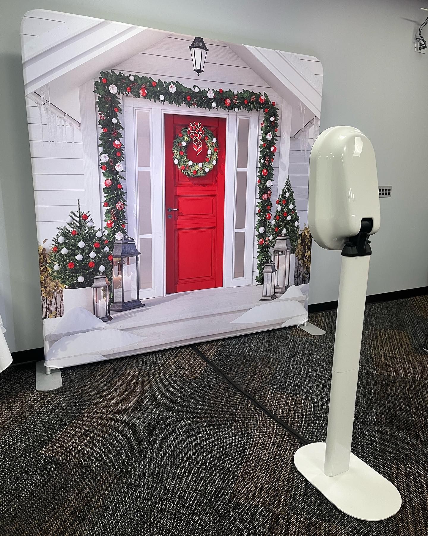 Photo booth with Christmas-themed backdrop: a red door with wreath, festive greenery, and a white, upright camera.
