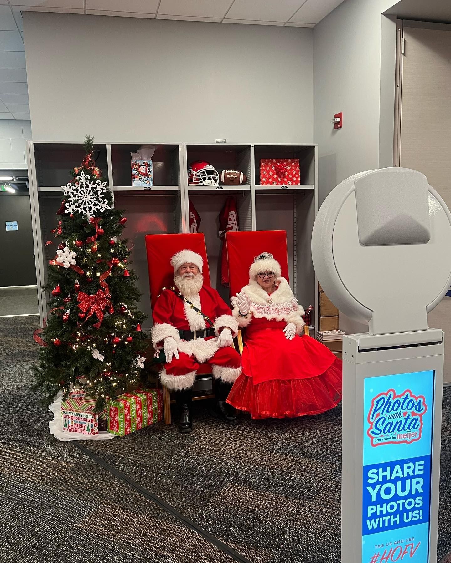 Santa and Mrs. Claus sit in chairs next to a Christmas tree and photo booth. They smile, posing for photos.