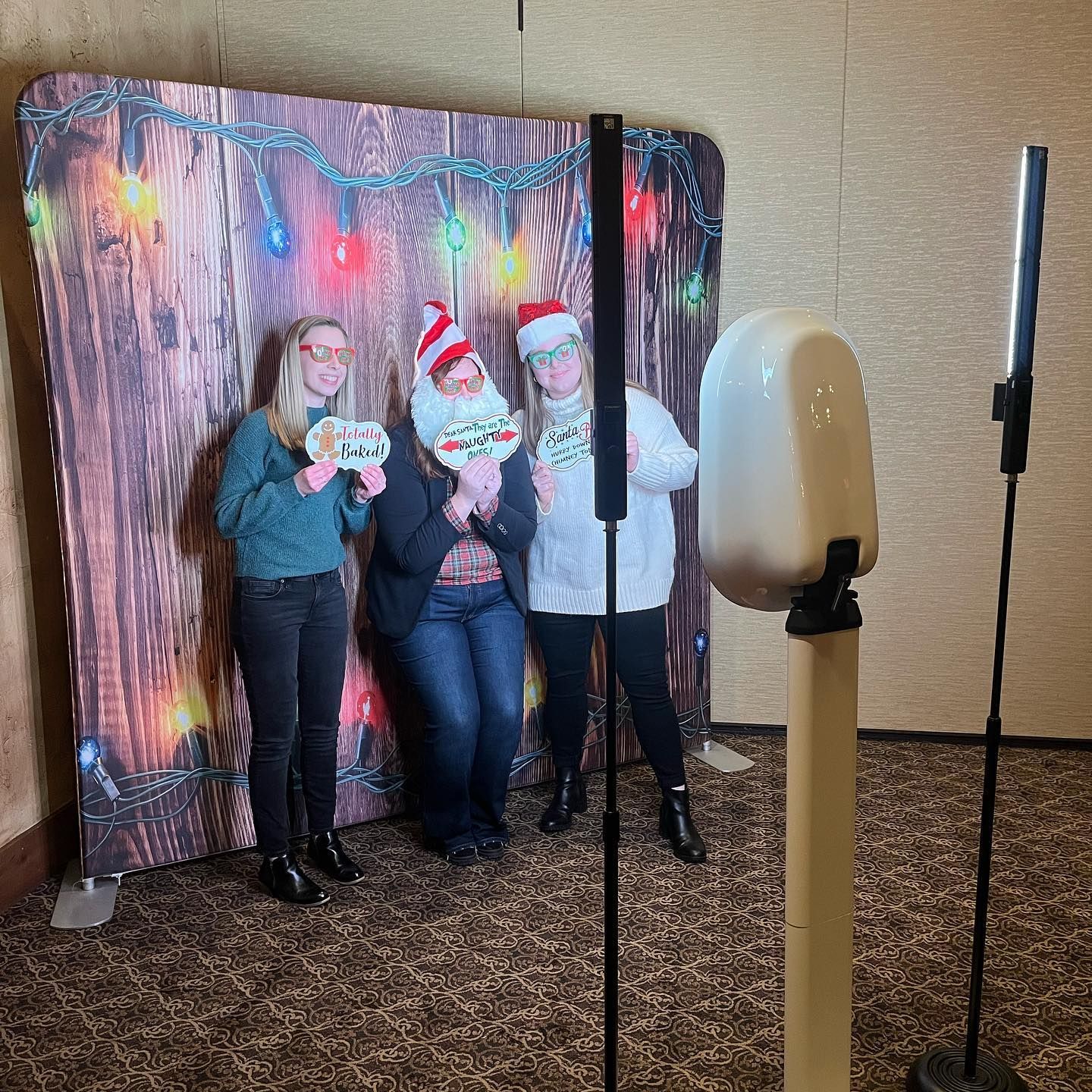Three people pose in front of a Christmas photo booth. They hold props and smile. A camera and lights are set up.