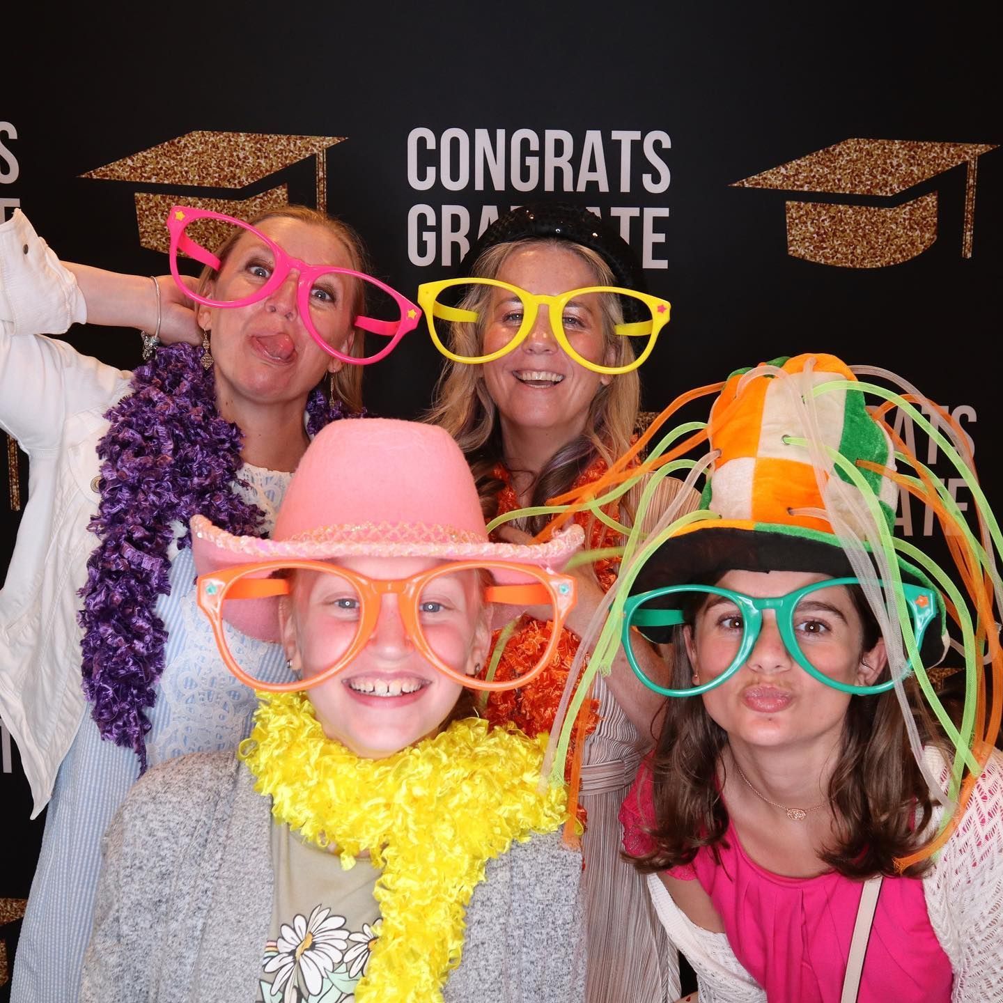 Four people wearing silly glasses and accessories pose in front of a graduation backdrop, making silly faces.