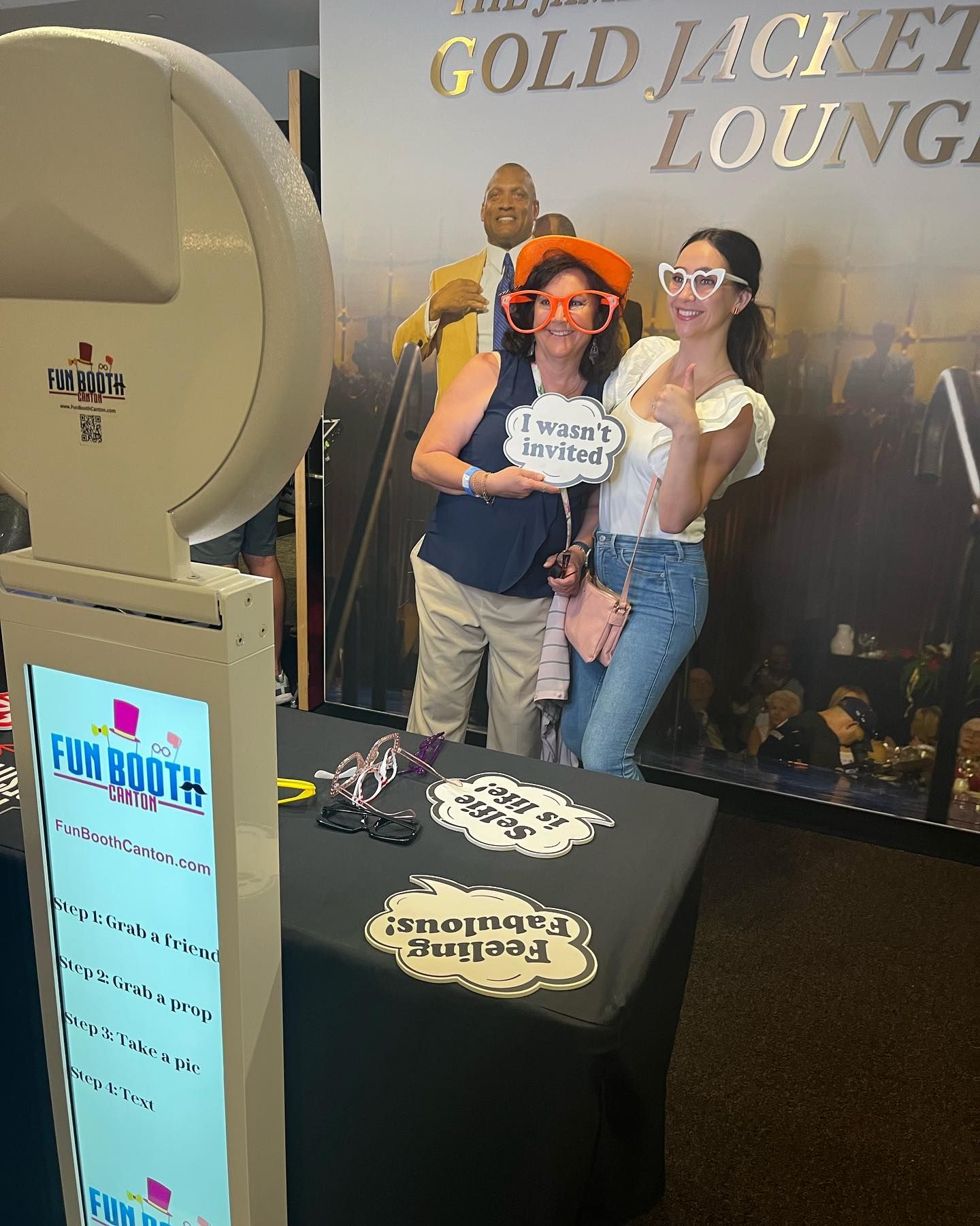Two women pose at a photo booth; one holds a sign, wearing novelty glasses. Black table with props.
