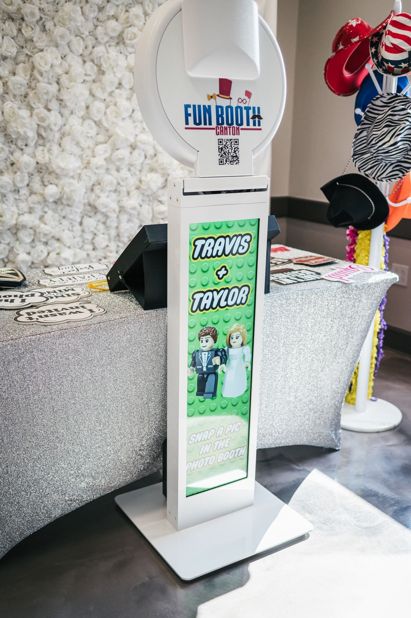 A white photo booth with a screen displaying a pixelated wedding couple. Hats and props are in the background.