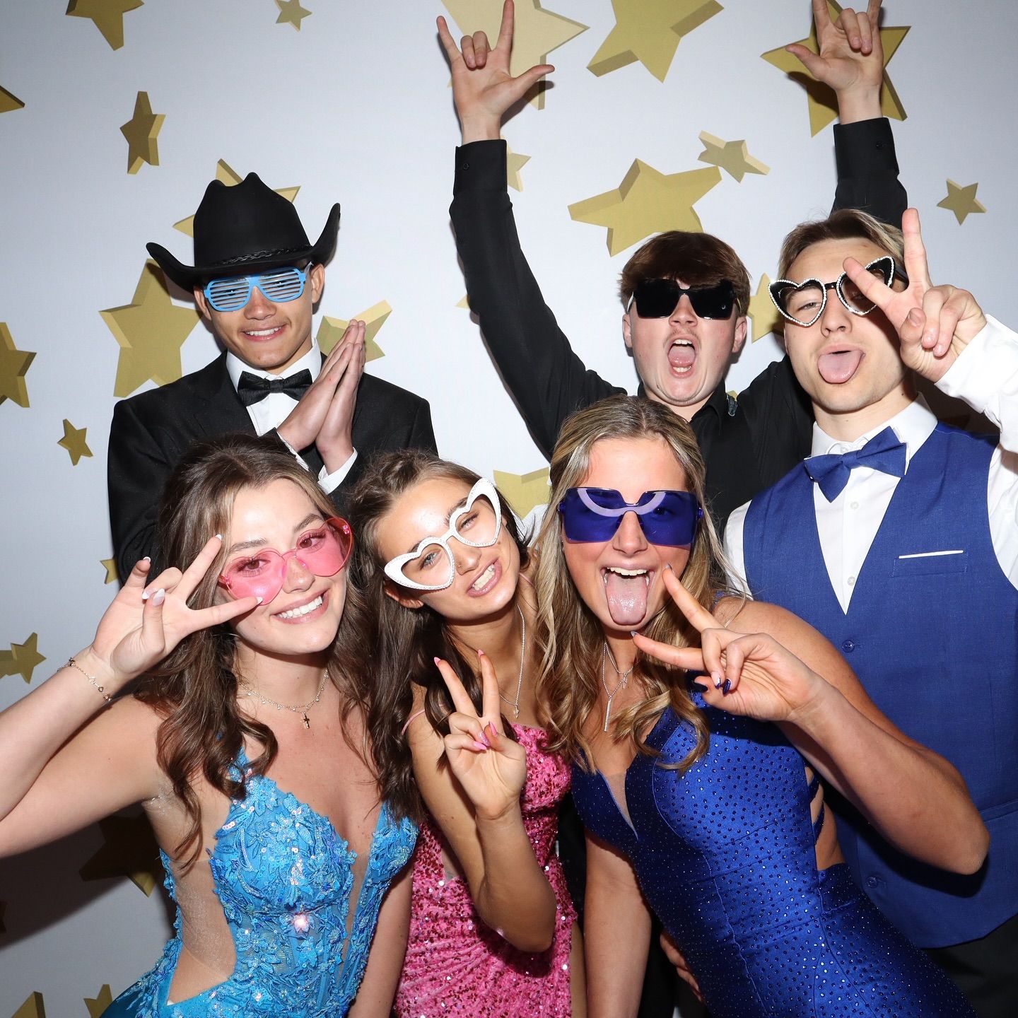 Six people in formal attire pose for a photo. Some wear novelty glasses and make hand gestures against a gold star backdrop.