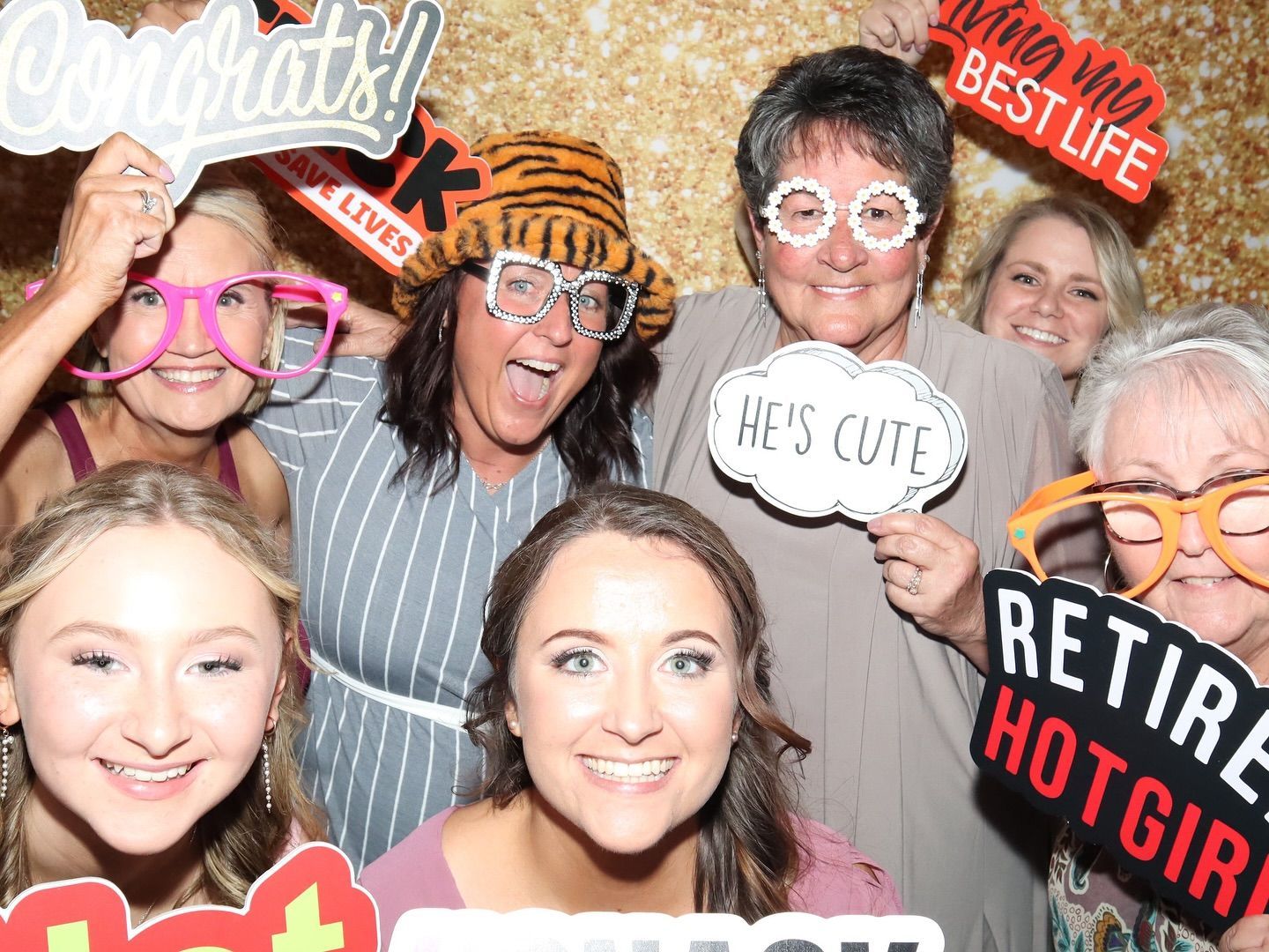 Group of smiling people posing with photo booth props against a gold backdrop.