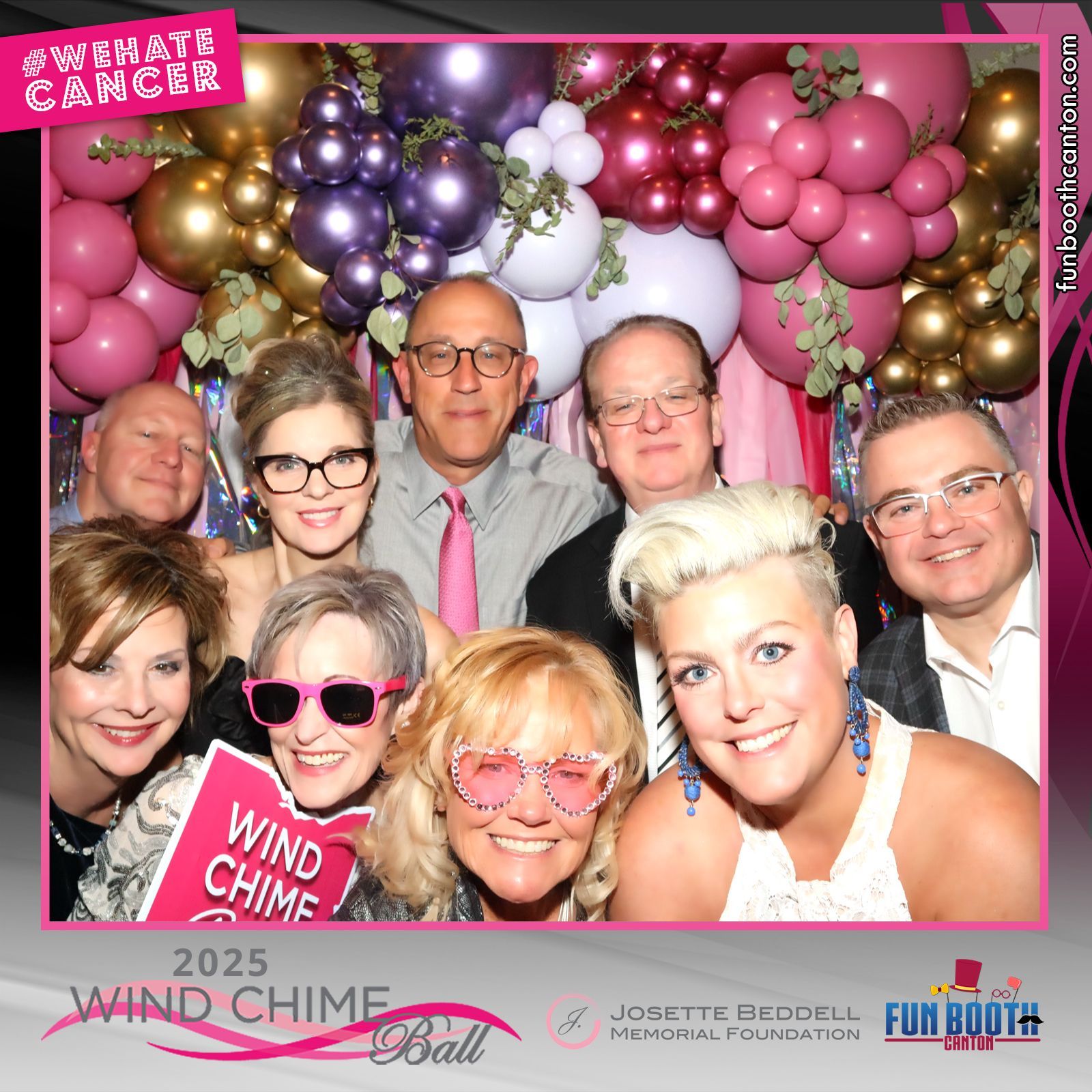 People pose at a Wind Chime Ball photo booth, smiling among pink, purple, and gold balloons.