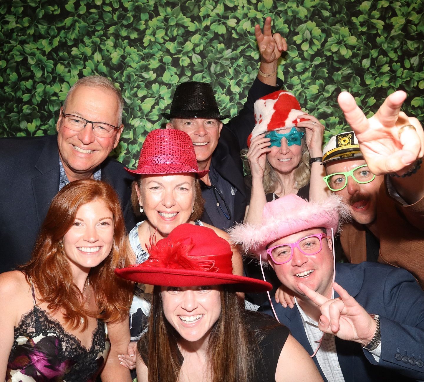 Group of people smiling, wearing hats, posing for a photo booth picture. Green leafy background.