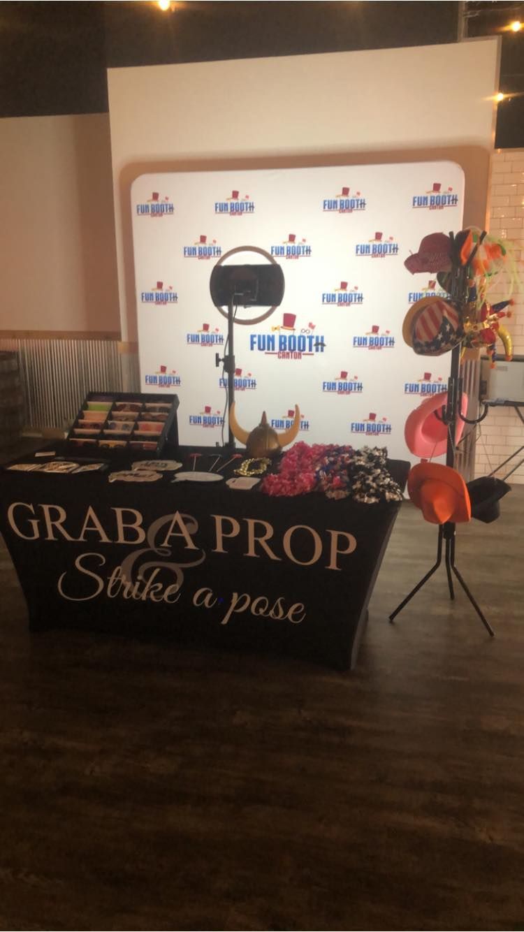 Photo booth setup with props on a table, backdrop, and stand with hats. Text says 