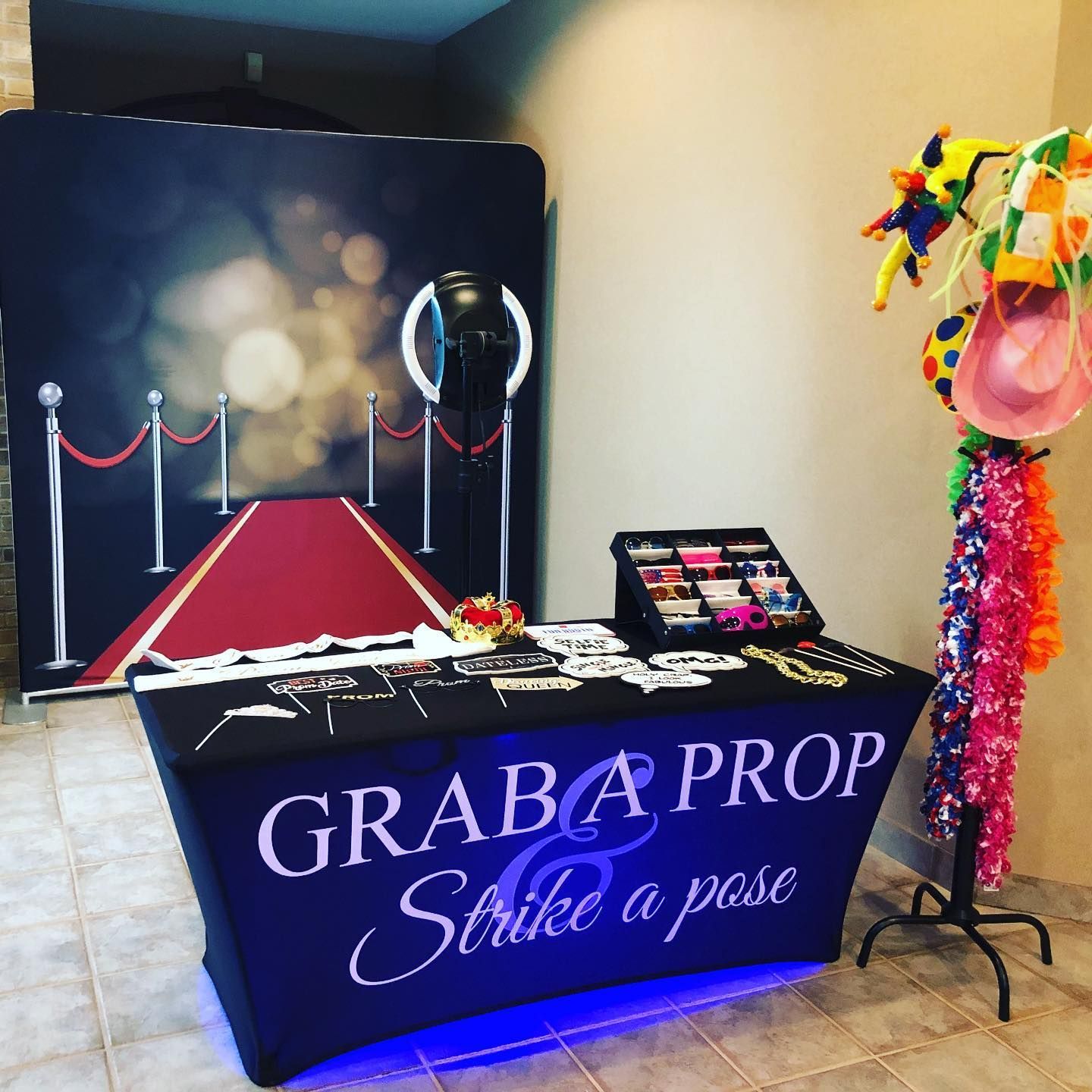 Photo booth setup with red carpet backdrop, props on a table, and a ring light. The table says 