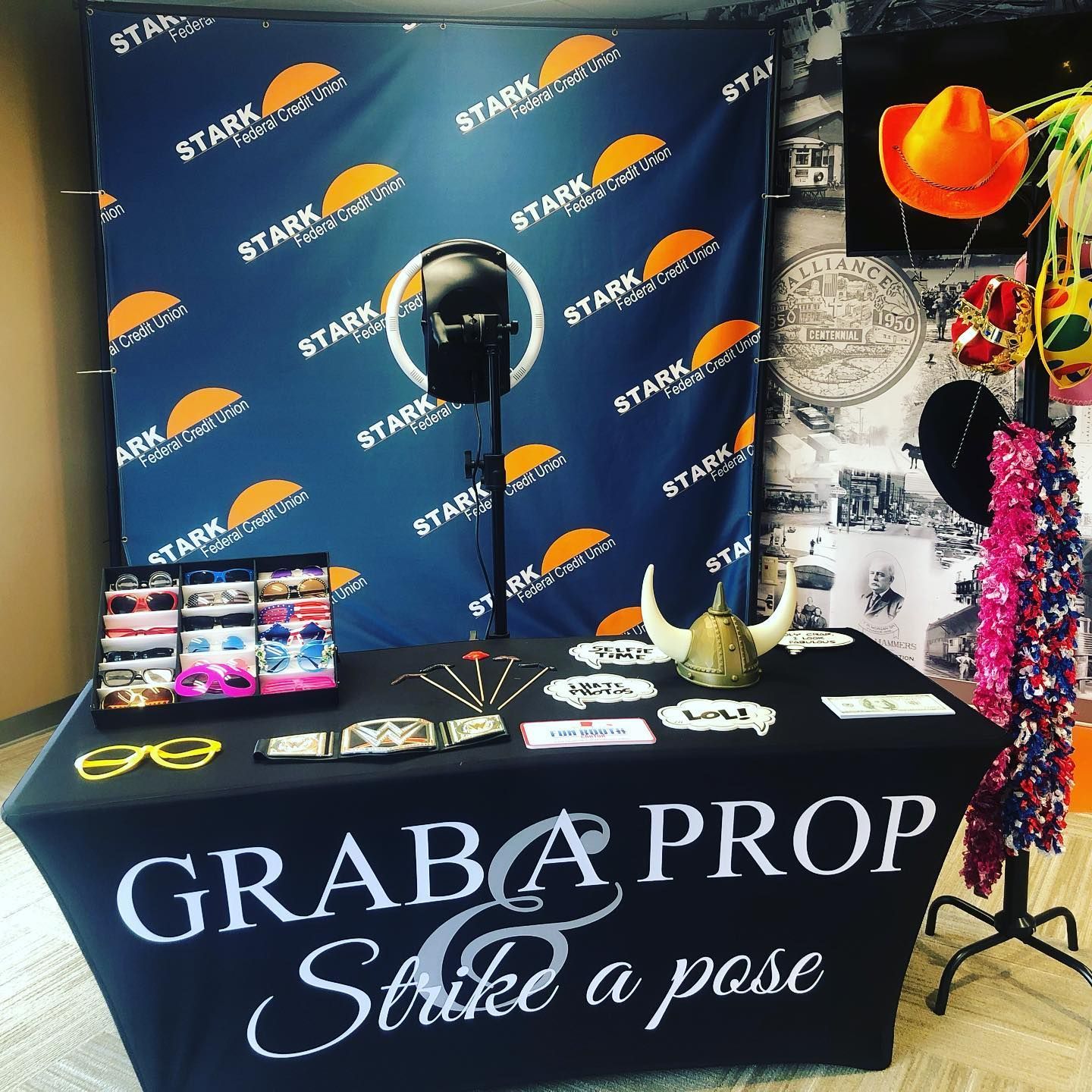 Photo booth setup with props on a table. Backdrop has a logo. Props include glasses, signs, and a lei.