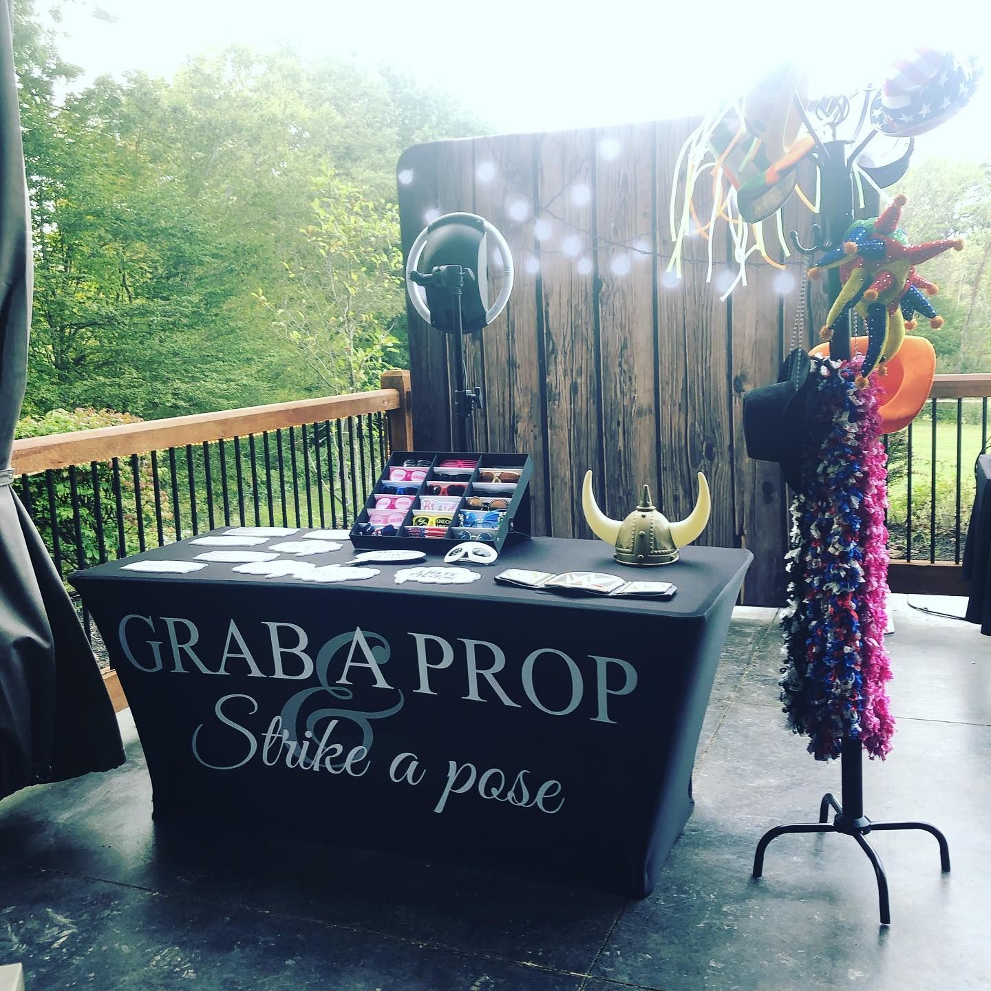 Photo booth setup on a deck: table with props, backdrop, mirror, hats, leis, and a sign that says 