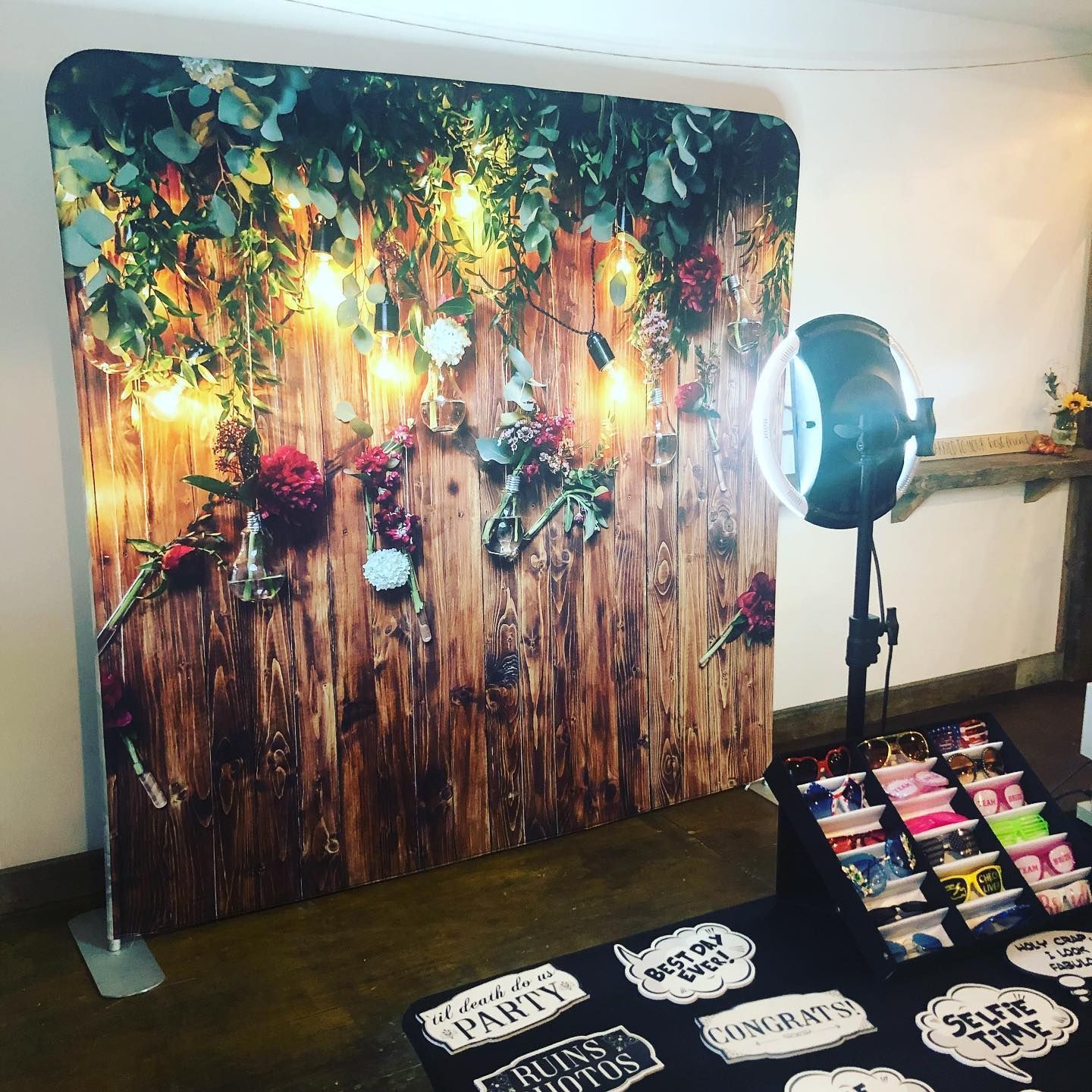 Photo booth setup: backdrop of wood and flowers, ring light, props on table.