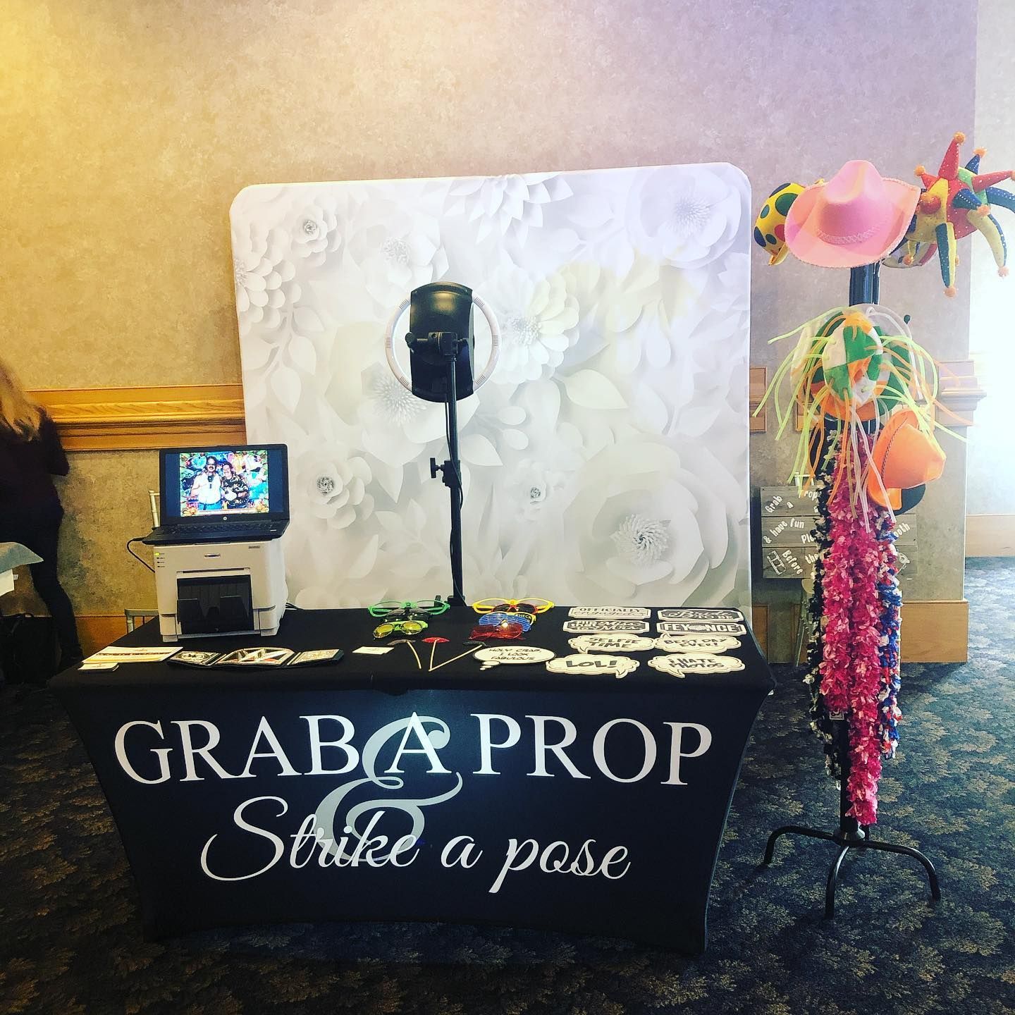 Photo booth setup: white flower backdrop, props, camera, printer, and sign that reads “Grab a prop & Strike a pose.”