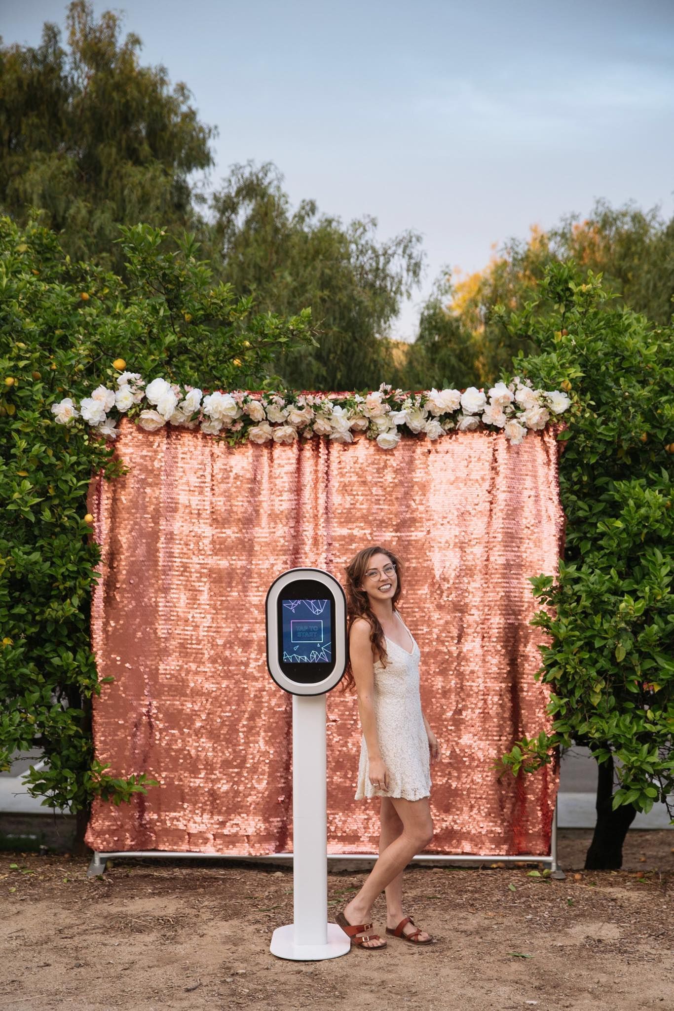 Woman smiling near a photo booth with rose gold sequin backdrop, outside with trees.
