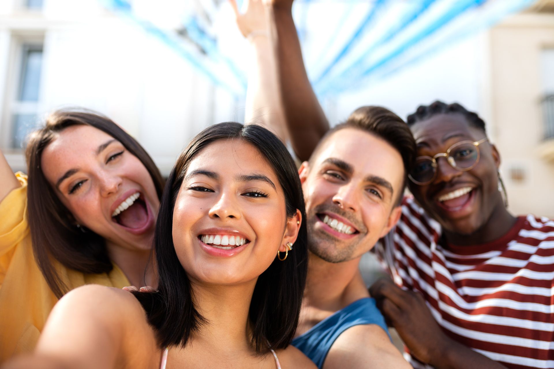 Four smiling people taking a selfie outdoors, arms raised. Bright sunlight, blue and white background.