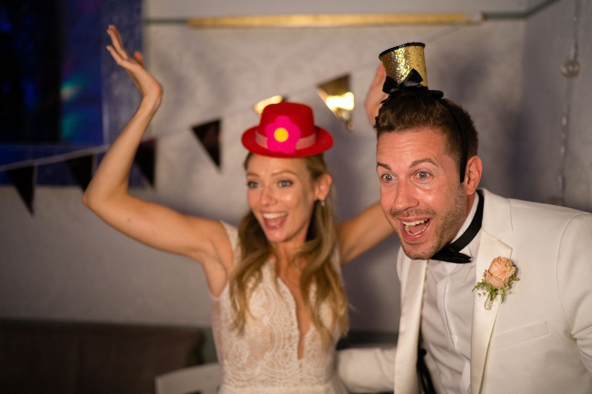 Newlyweds with party hats celebrating. Bride in white dress, groom in white suit, both excited.