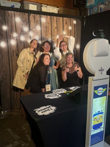 Five people pose in front of a photo booth with a wood backdrop and string lights; two are giving thumbs up.