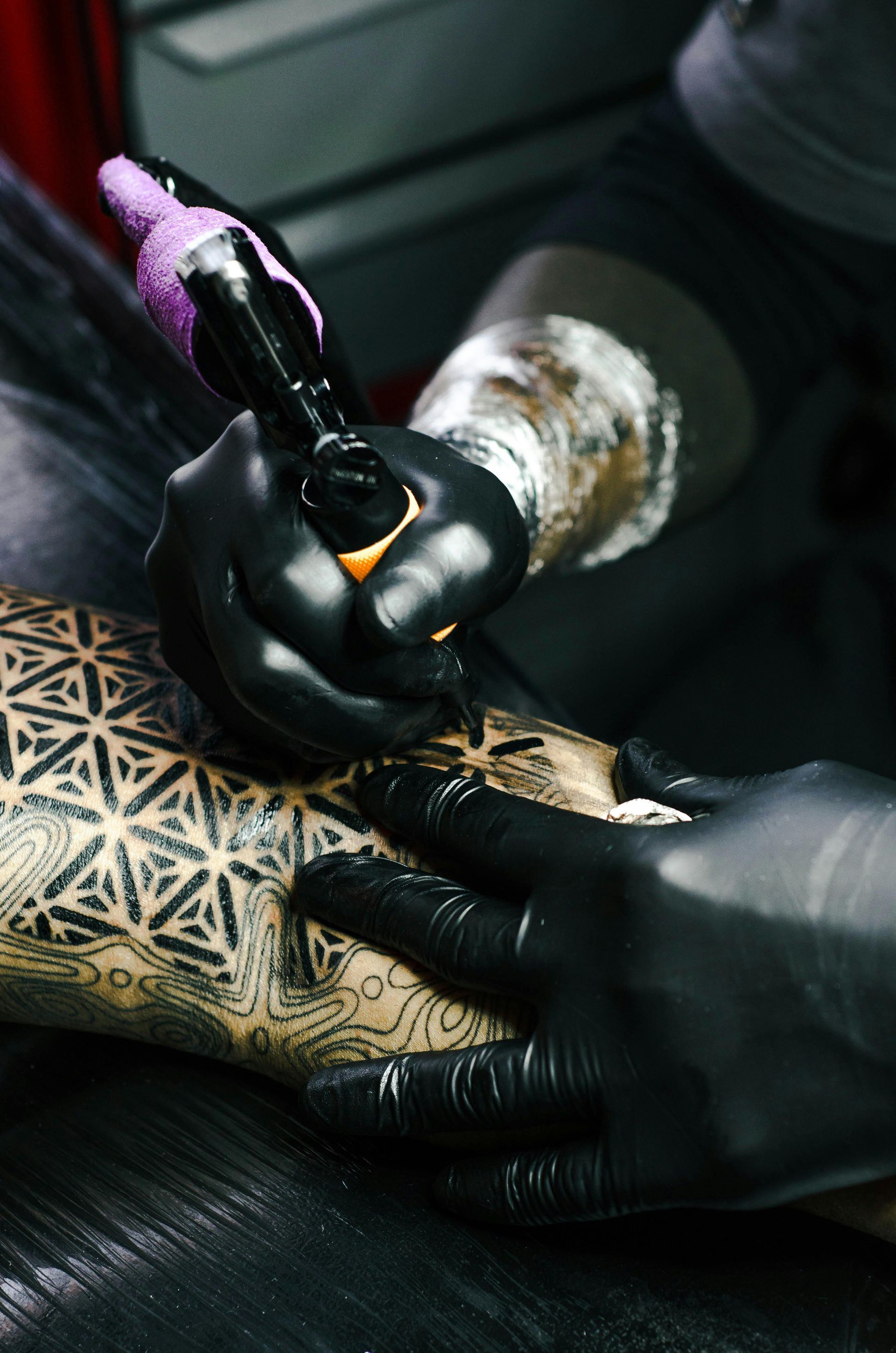 Tattoo artist wearing black gloves inks geometric pattern on a person's skin.