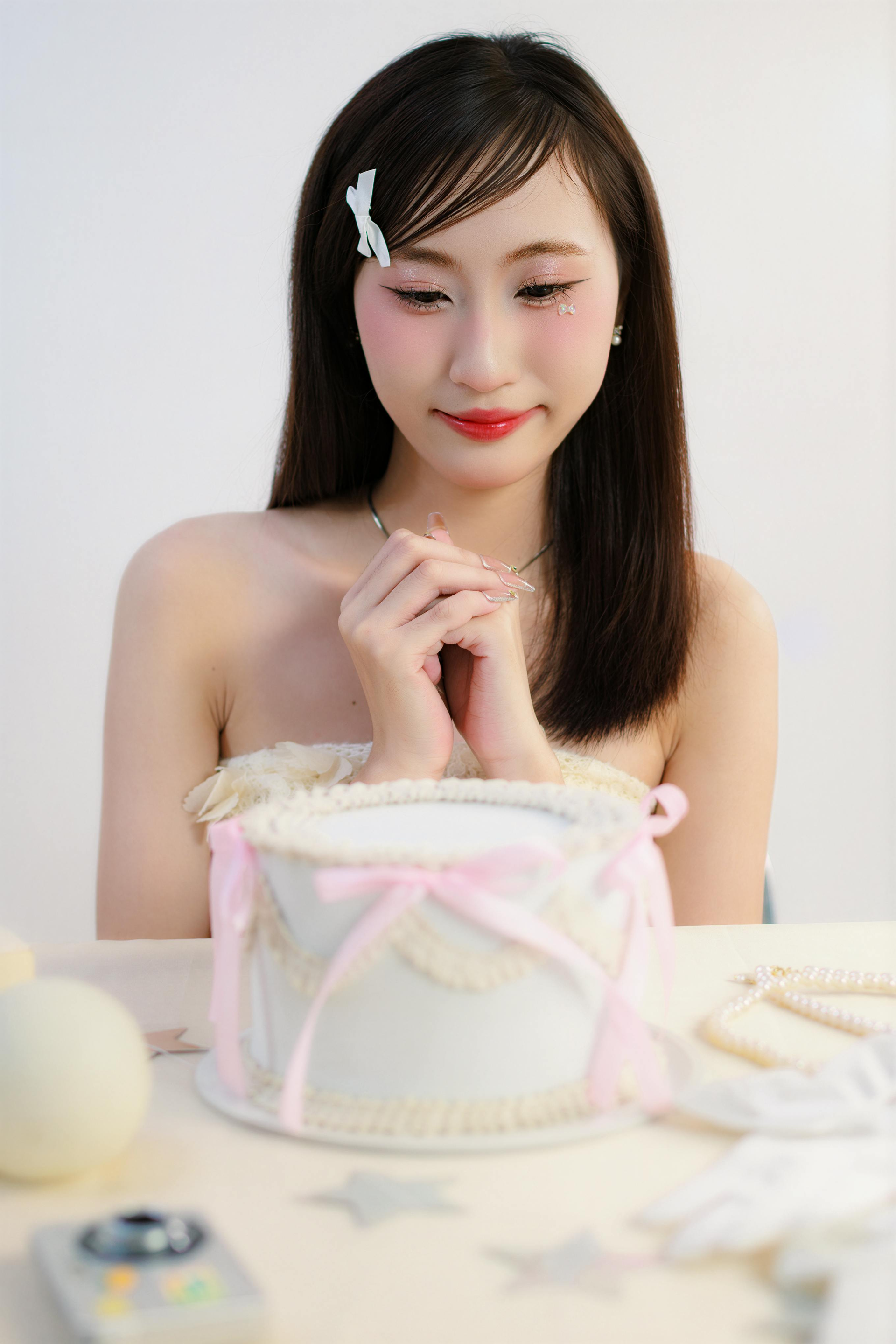 Woman with pink blush and red lipstick, looking at a cake with pink ribbons on a table.