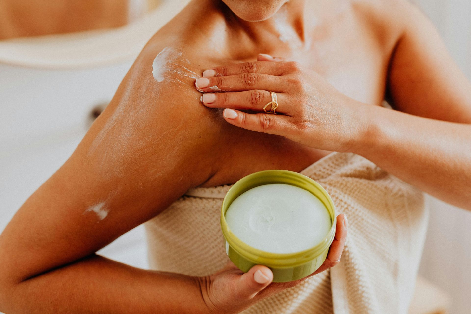 Woman applying cream to her shoulder from a green jar, wrapped in a towel.