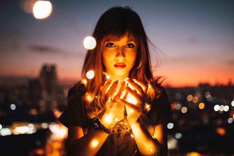 Woman holds string lights, glowing against a city sunset.