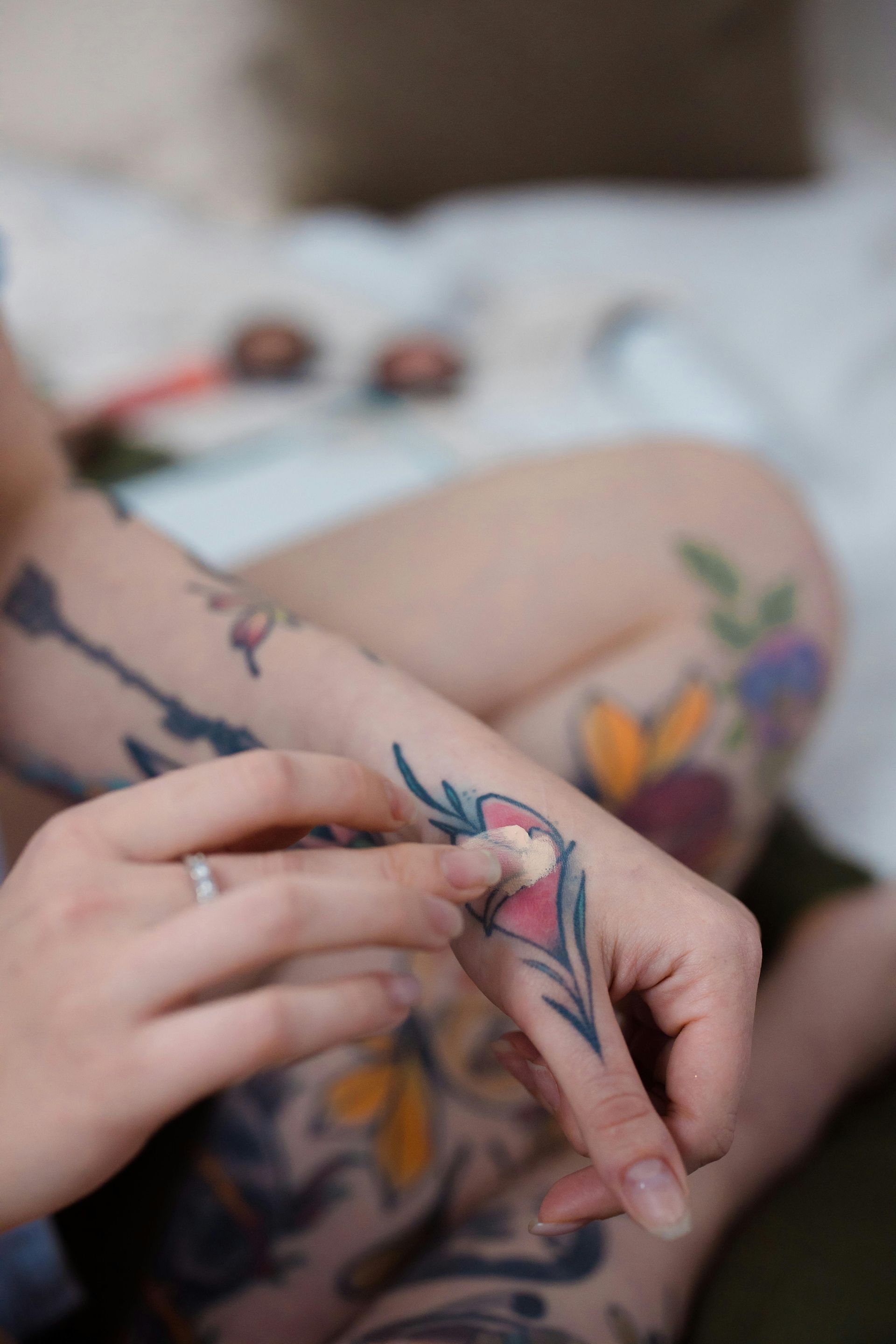 Person applying cream to a hand tattoo, surrounded by other tattoos, with a blurred background.