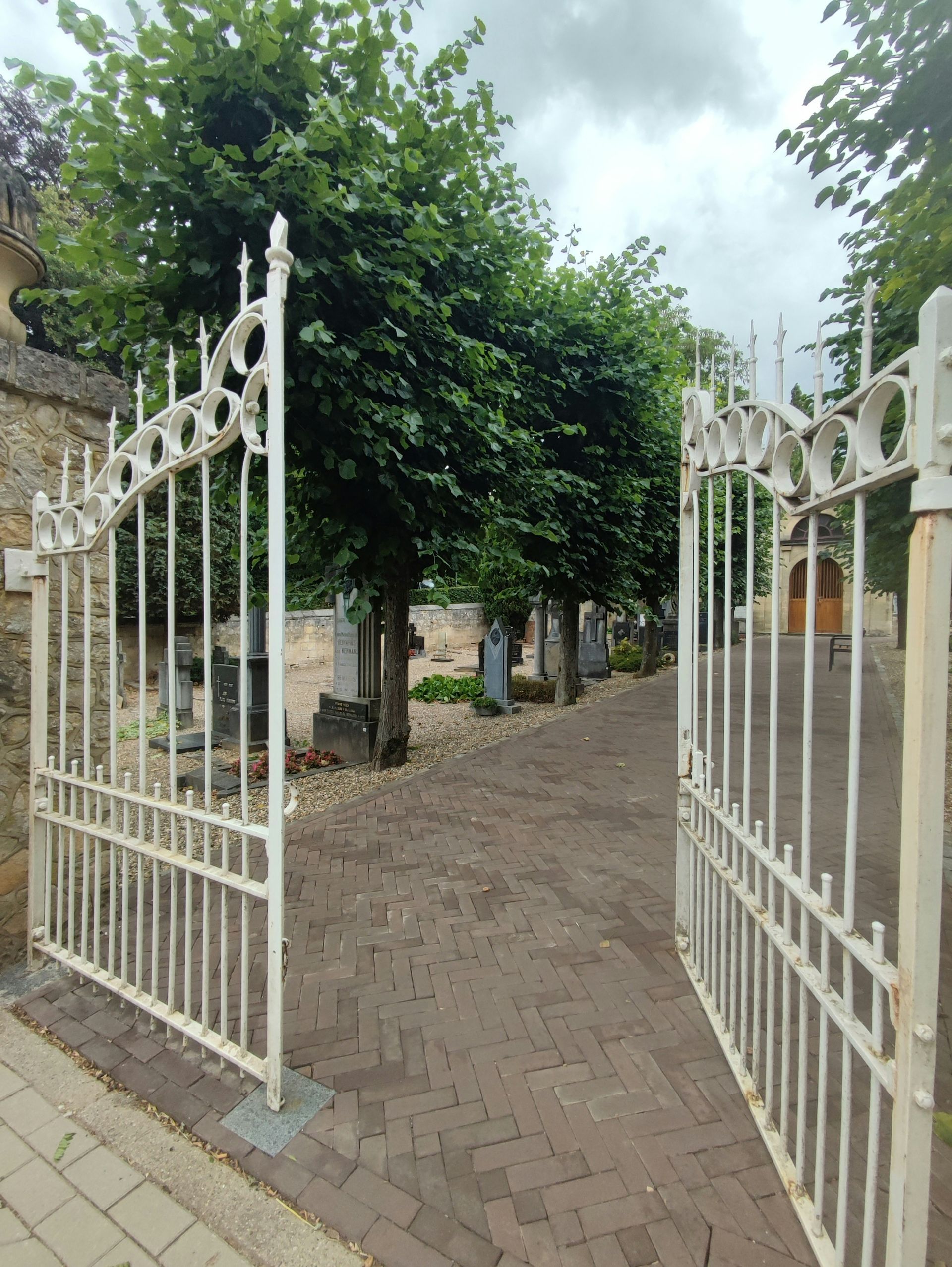 Open white wrought iron gates leading into a cemetery with headstones and trees. Brick path.