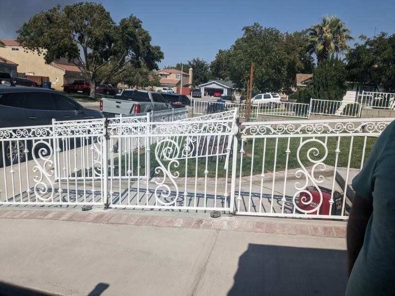 White ornate metal fence bordering a residential front yard with cars and houses in the background on a sunny day.