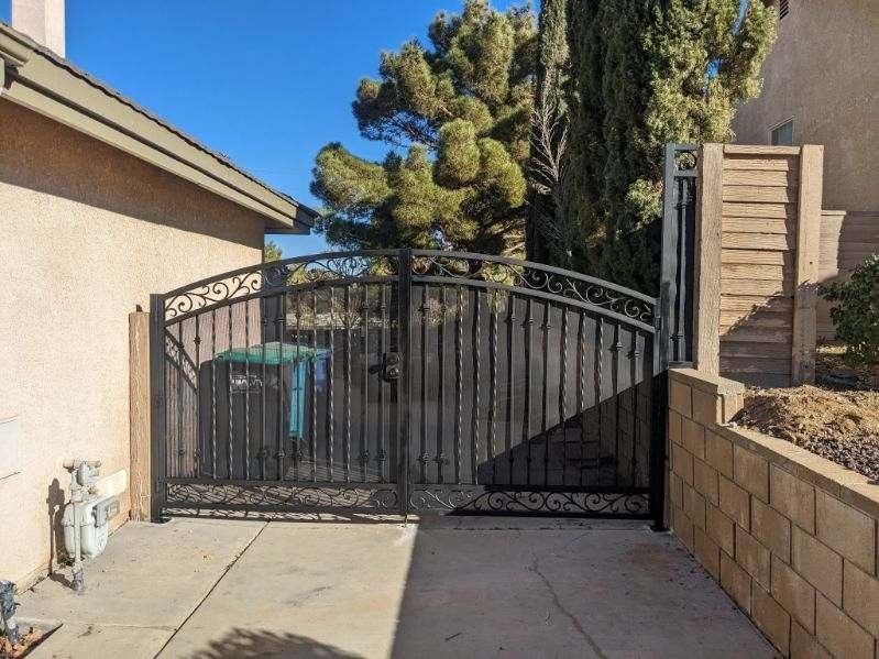 Black wrought iron gate on a concrete driveway, next to a beige house and retaining wall, under a blue sky.