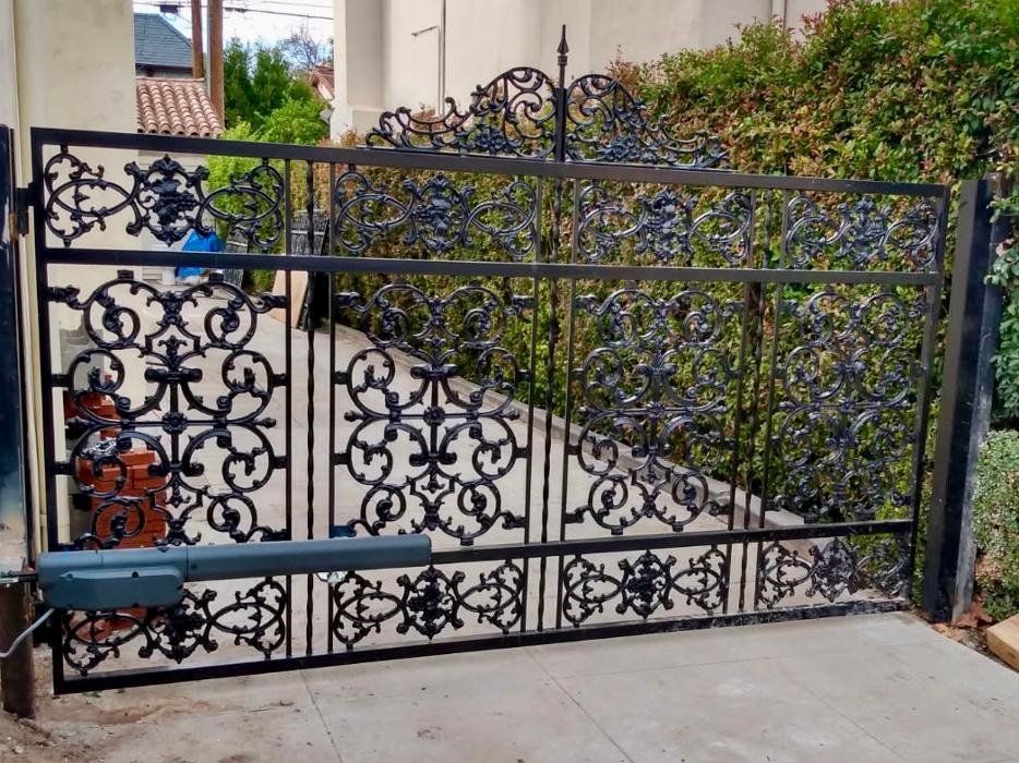 Black wrought iron gate with decorative scrollwork, automated opening, in front of a green hedge.