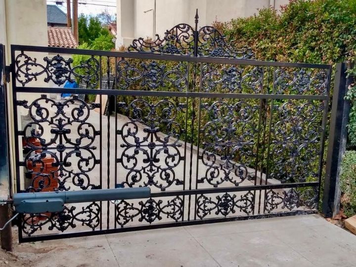 Black wrought iron gate with ornate scrollwork, automated with a motor, set against a green hedge.
