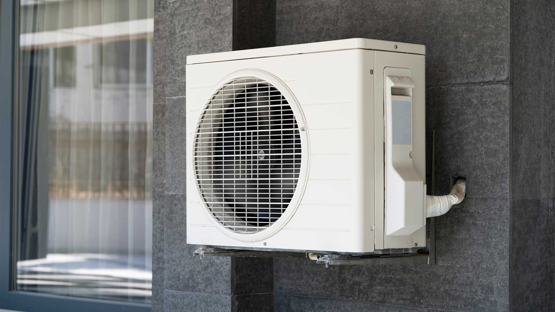 A white air conditioner is mounted on the side of a building next to a window.