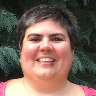 Woman with short dark hair smiles in front of green foliage; wearing a pink top.