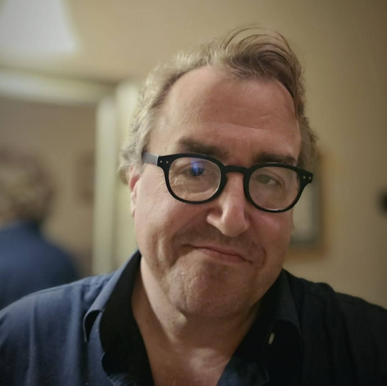 Man with glasses smiles, looking at the camera. He wears a dark shirt indoors, a mirror behind him.