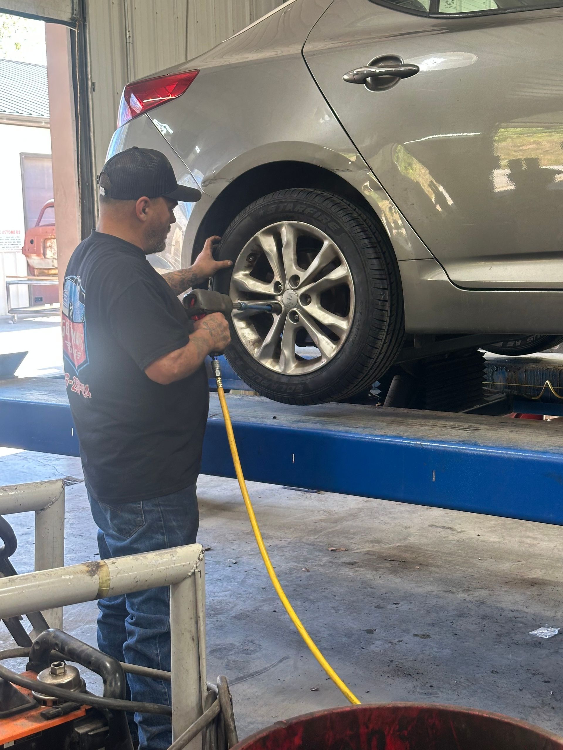 Mechanic using a tool on a car tire, in a garage setting with blue lift and a yellow hose. | Gentleman’s Garage