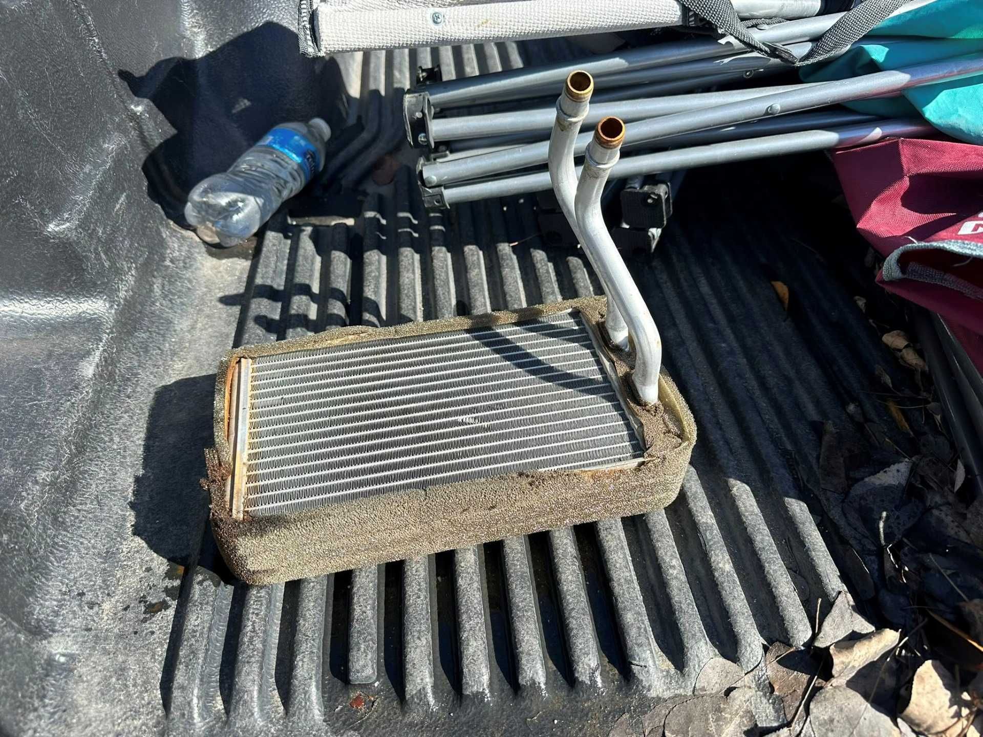 A heater core with two metal pipes rests on the ribbed metal floor of a truck bed next to a water bottle.