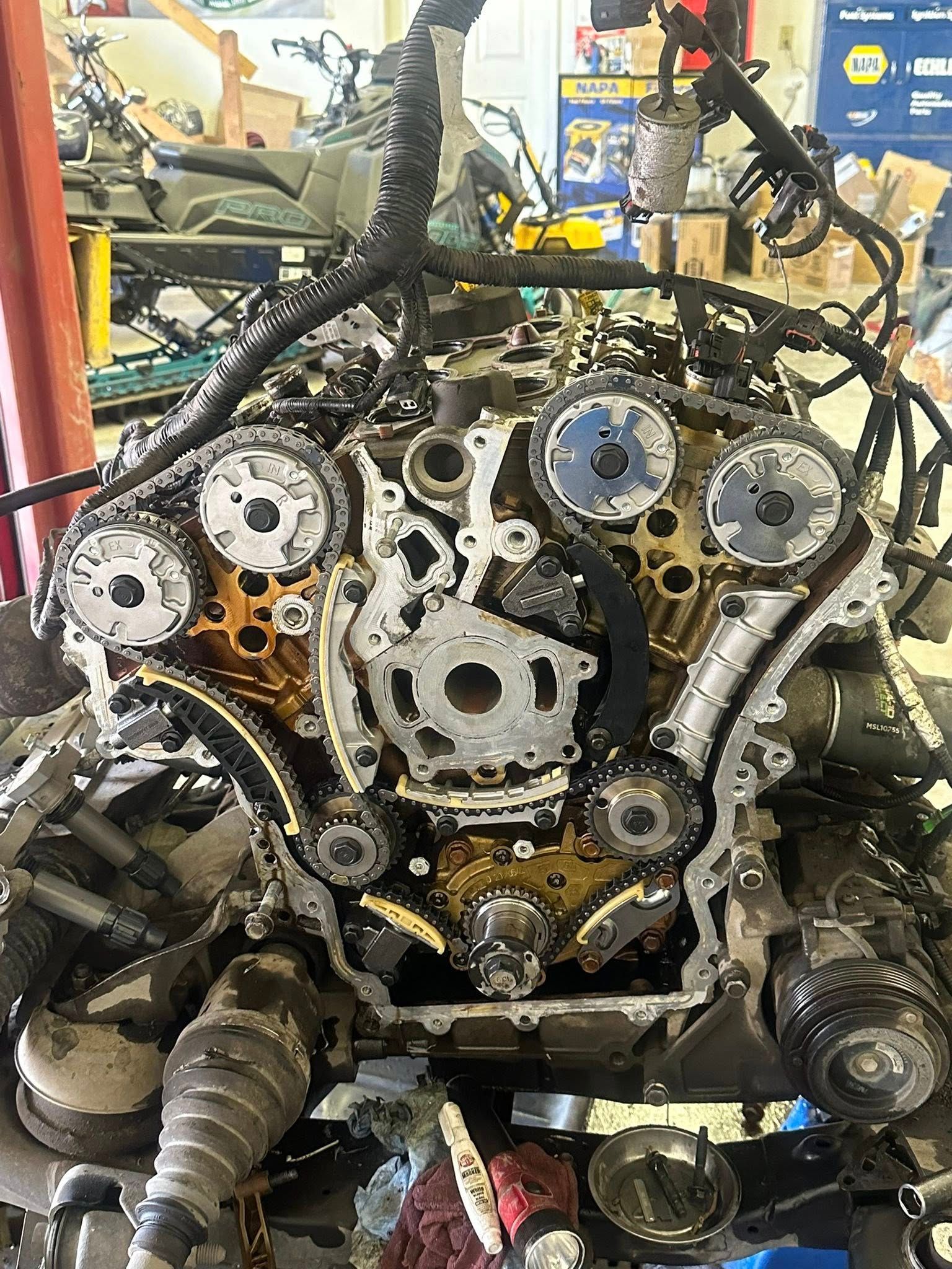 The open timing cover of a disassembled V6 engine showing gears, chains, and internal components inside an auto shop.