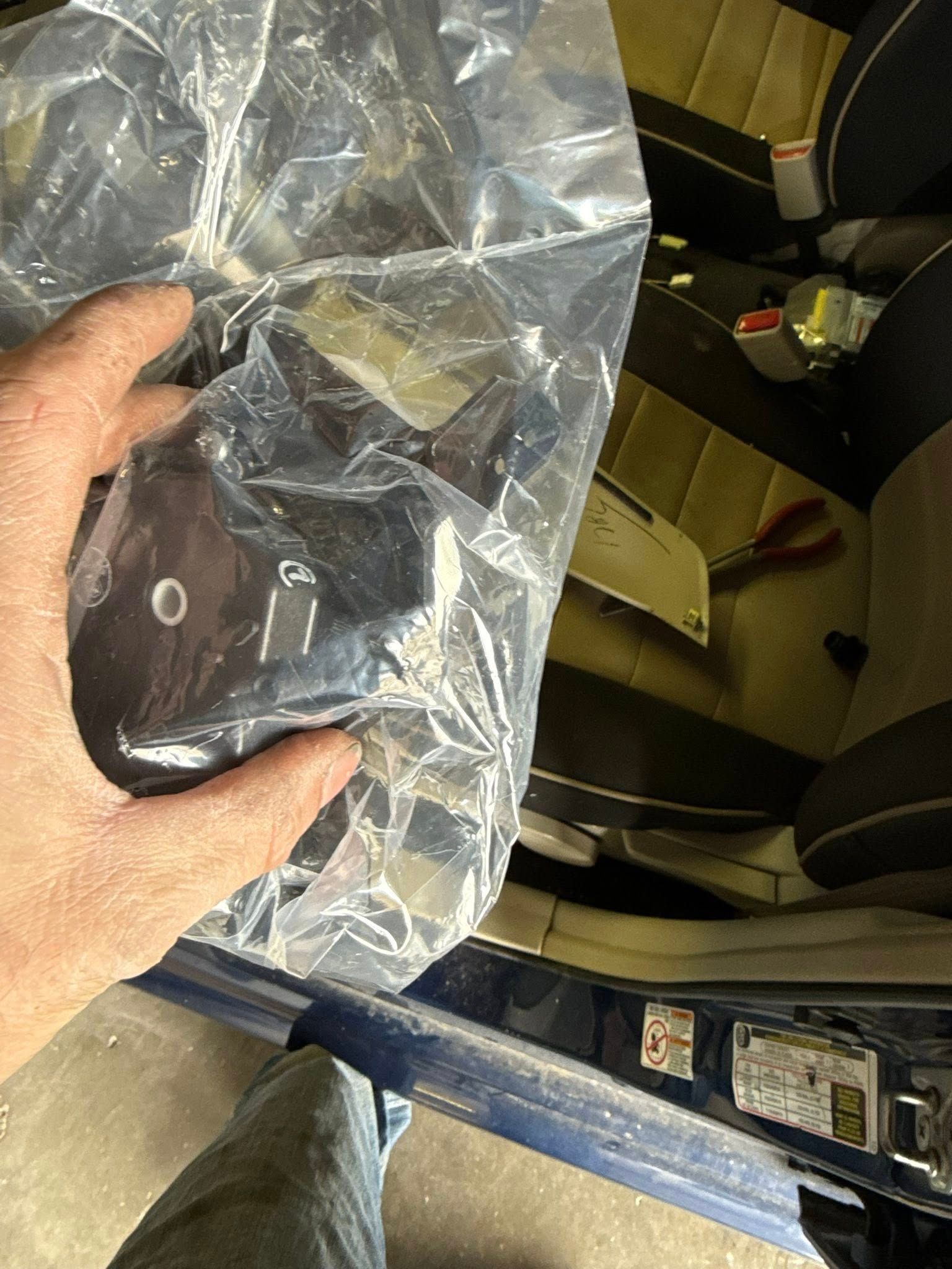 A hand holds a packaged black metal automotive part inside a car, with the vehicle's interior visible in the background.