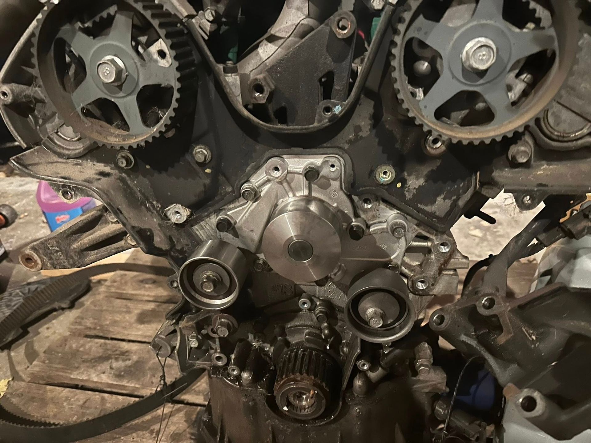 Car engine block front view showing timing pulleys, a central water pump, and a crankshaft gear.
