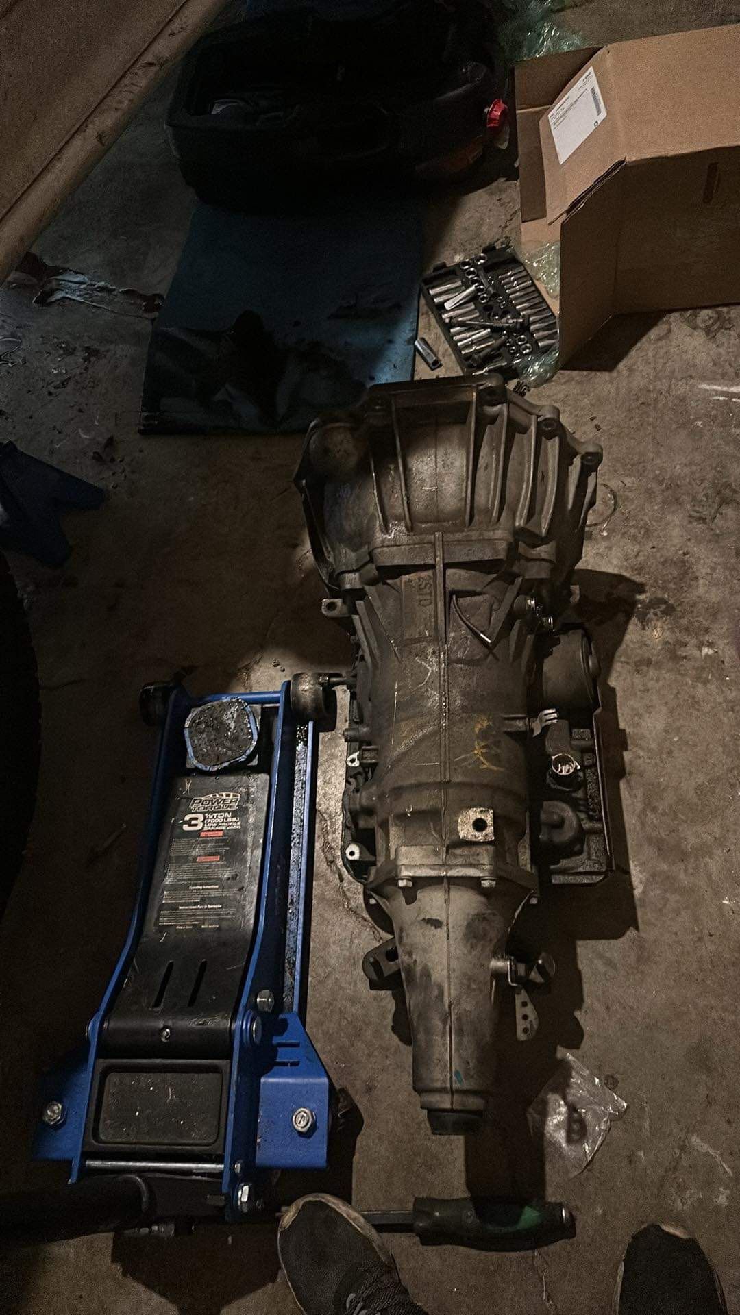 A car transmission sits on a garage floor next to a blue hydraulic floor jack and a cardboard box.