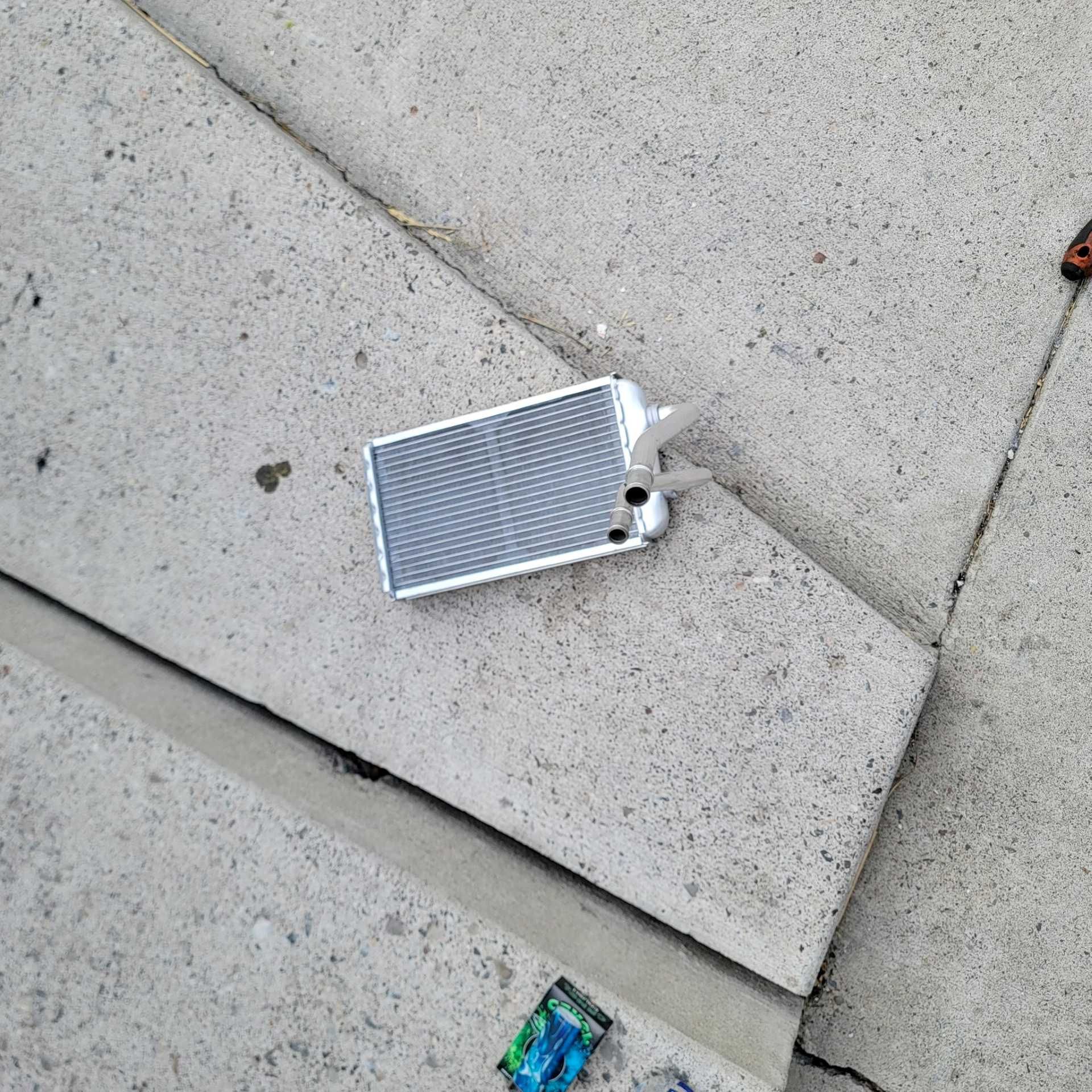 A small silver heater core radiator lying on a concrete surface, with a small colorful package nearby.