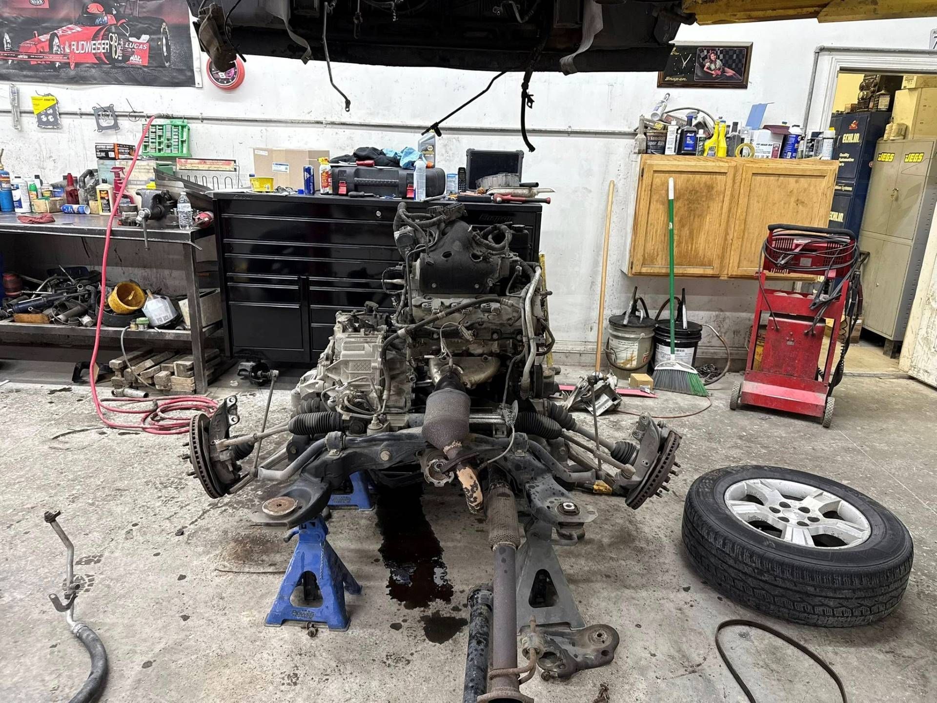 A vehicle engine and front chassis section supported by two blue jack stands in a messy automotive repair shop.