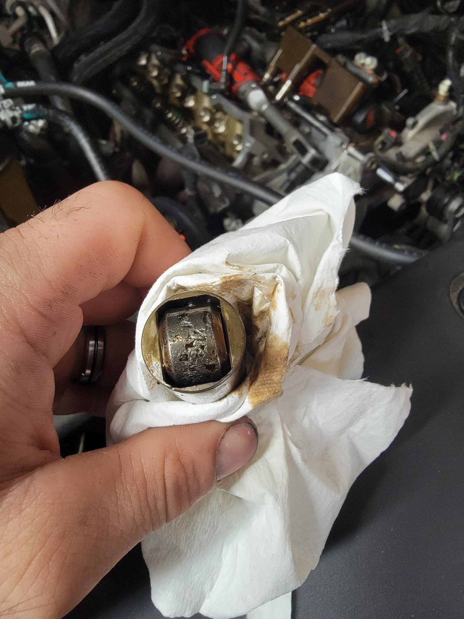 A hand holding a damaged automotive roller lifter covered in metal shavings against an engine background.