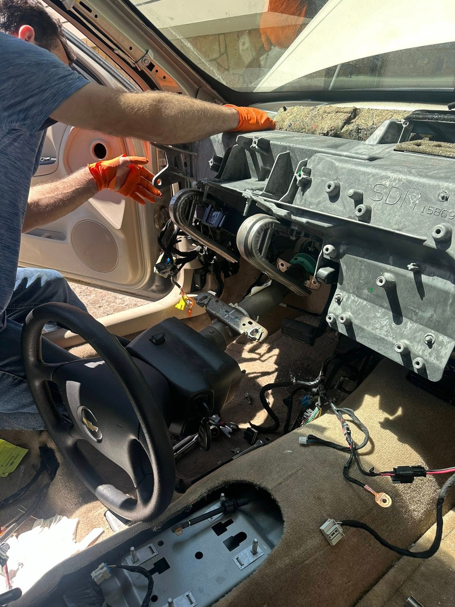 A person wearing orange gloves disassembles the dashboard of a vehicle, exposing the internal wires and structural frame.