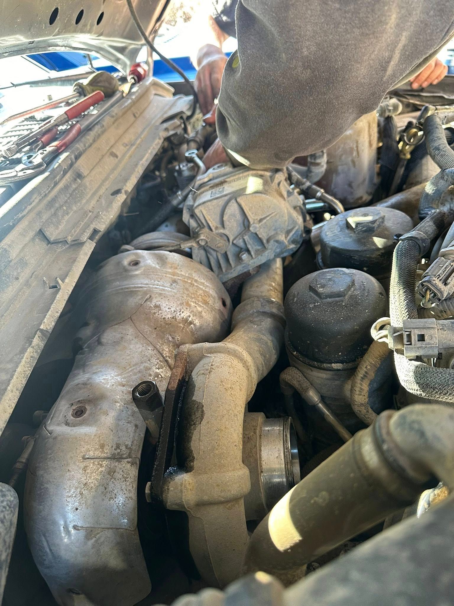 A person works on a dusty, metallic engine assembly inside an open vehicle hood.