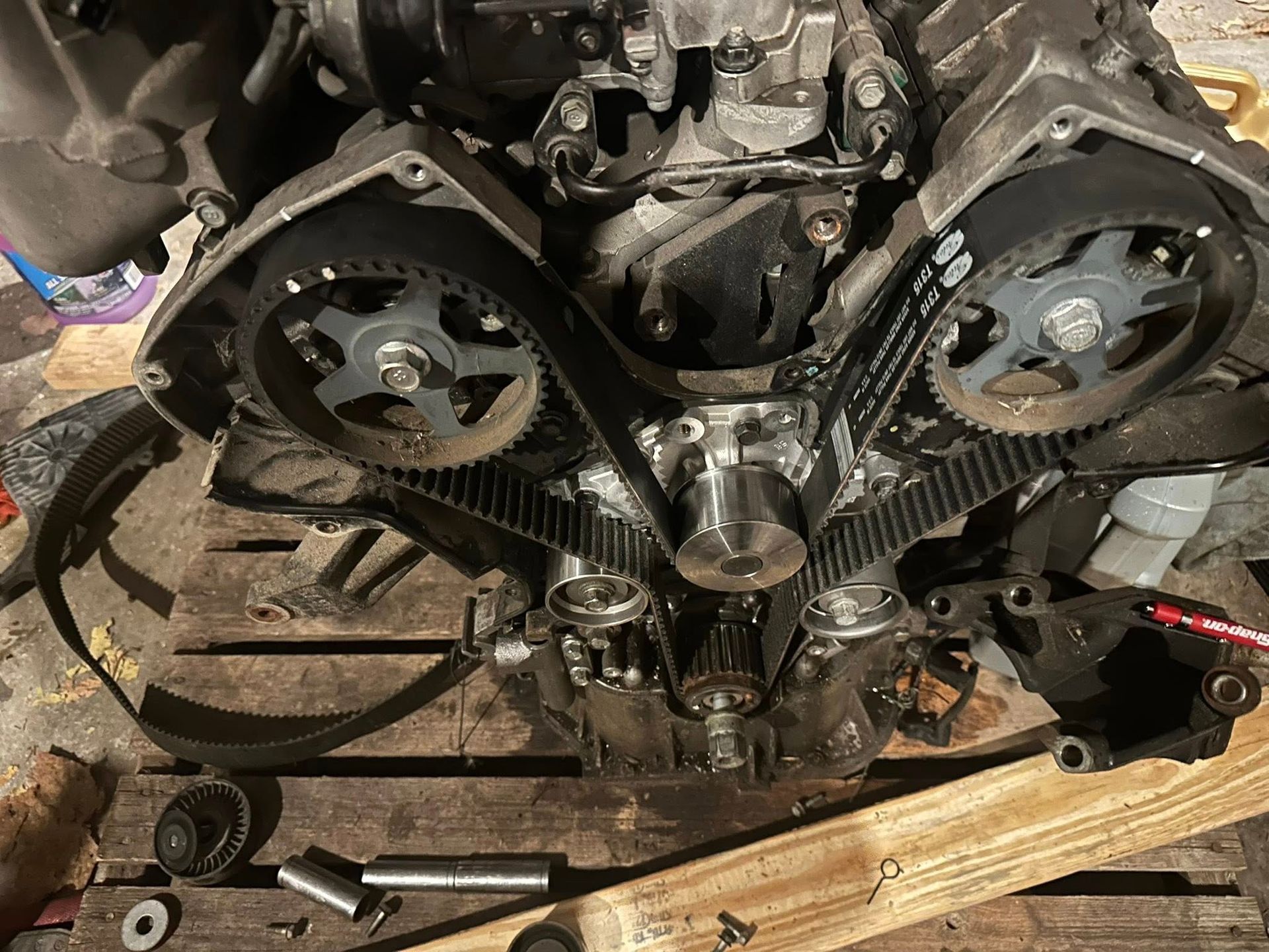 A timing belt installed on a disassembled car engine, featuring two camshaft pulleys and a crankshaft gear.