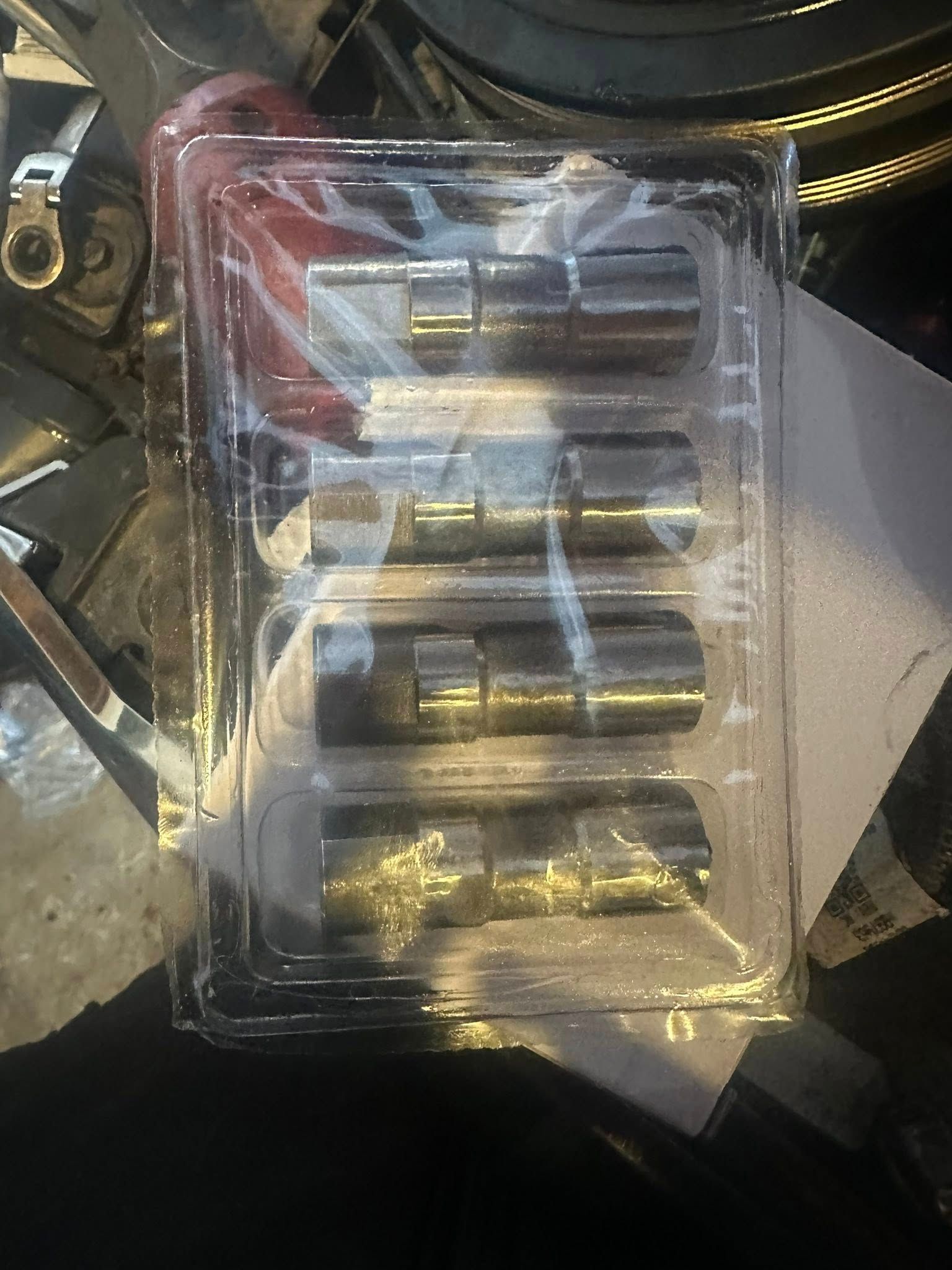 A clear plastic blister pack containing four cylindrical metal engine lifters resting on a cluttered workbench.