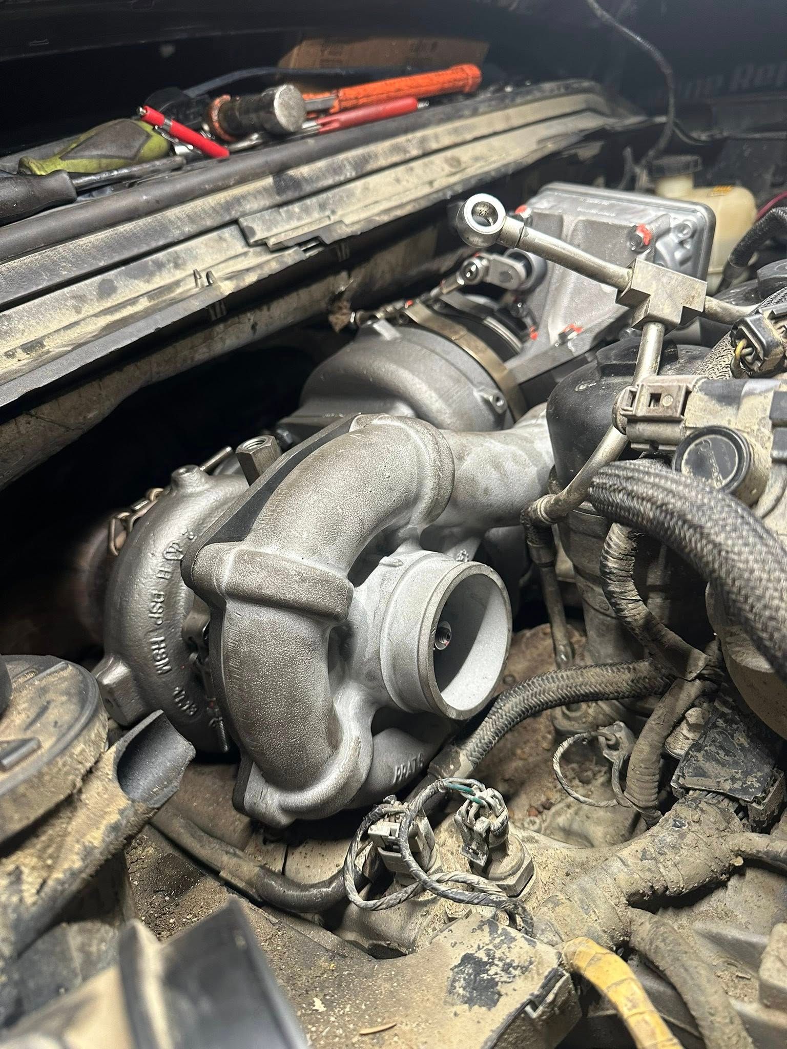 A close-up view of a dusty, silver automotive turbocharger installed in a vehicle engine bay.