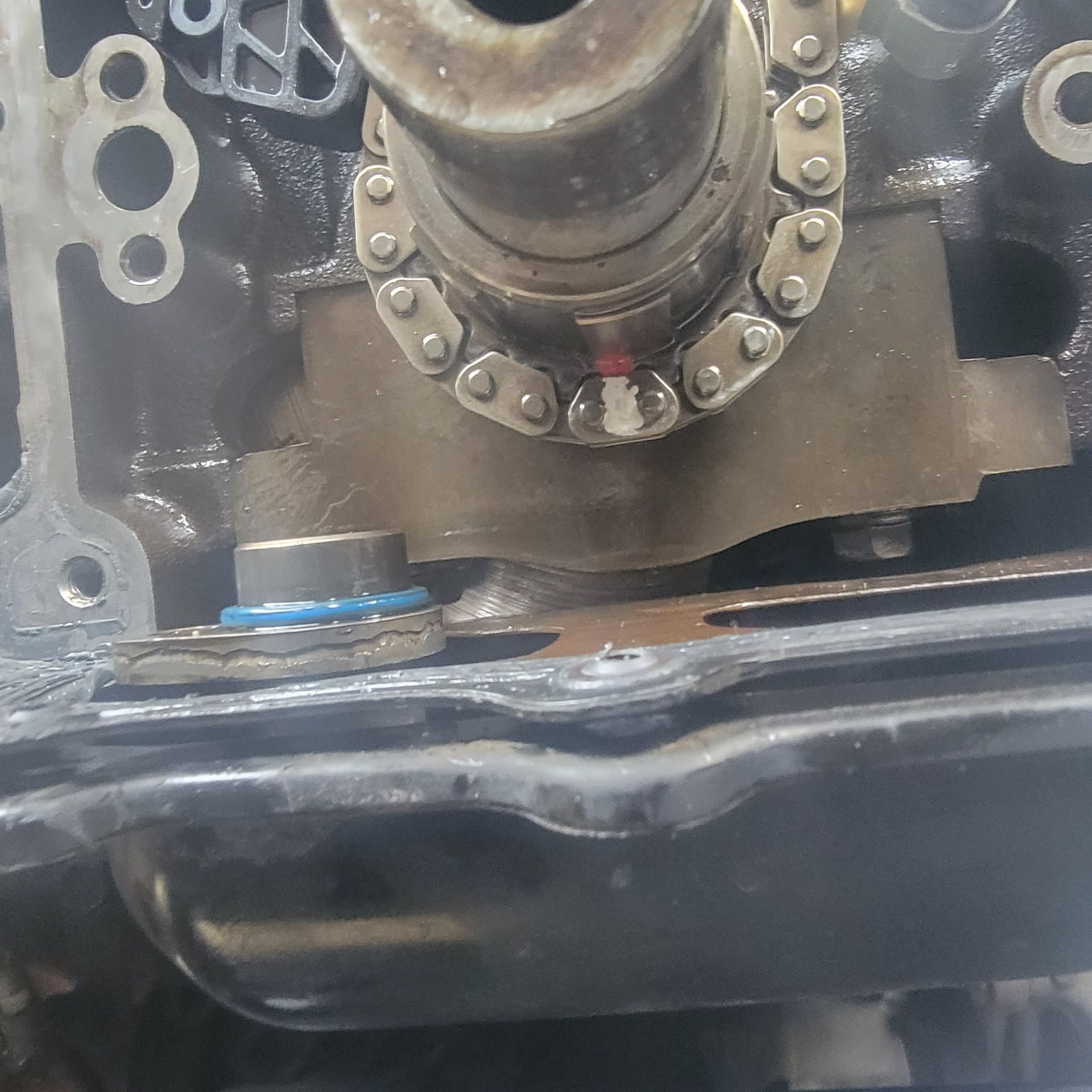A close-up of a vehicle engine block showing a timing chain gear, a oil pump pick-up tube with a blue seal, and oil pan.