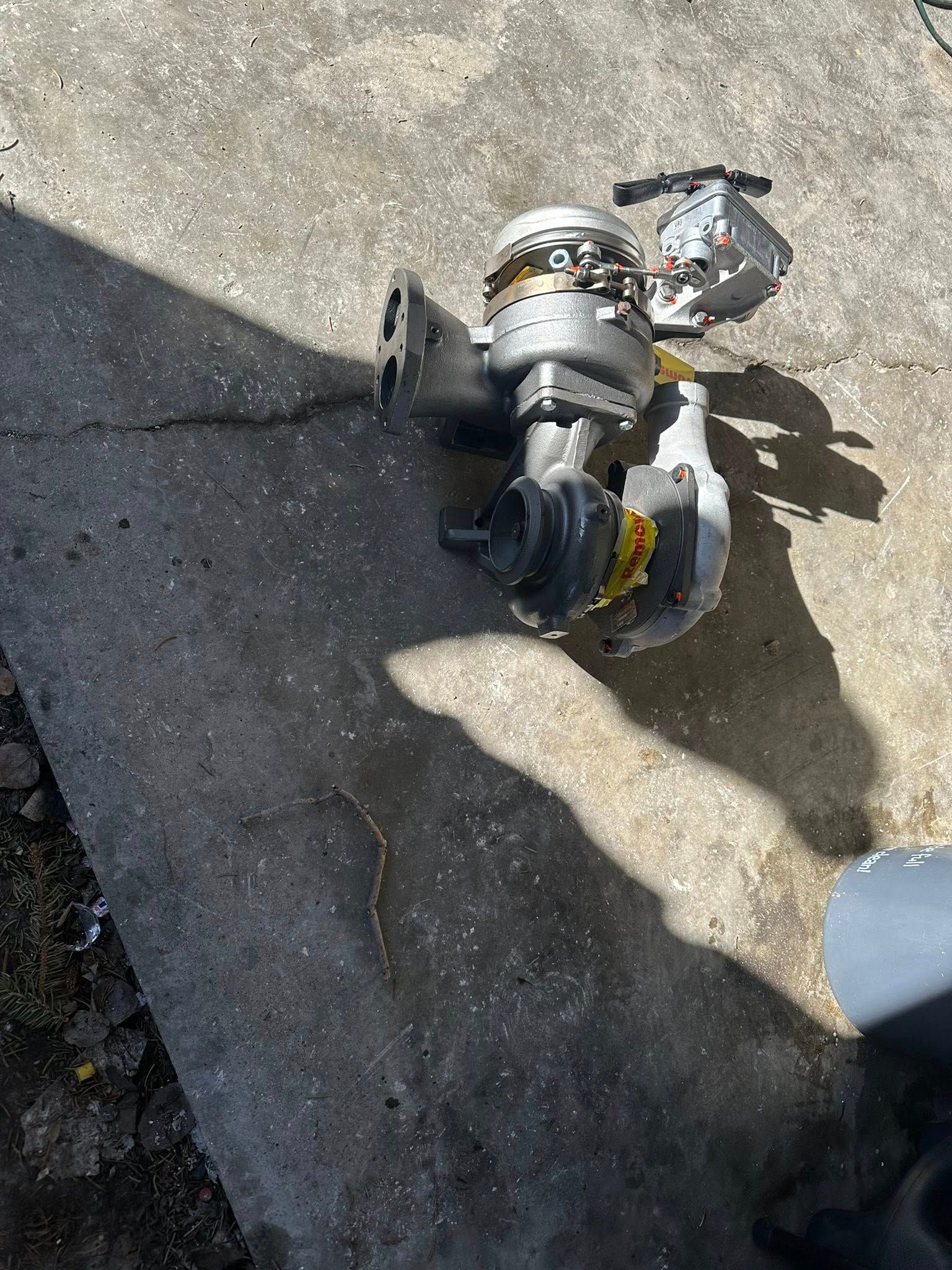A metal turbocharger unit sits on a concrete surface, casting a long shadow under bright sunlight.