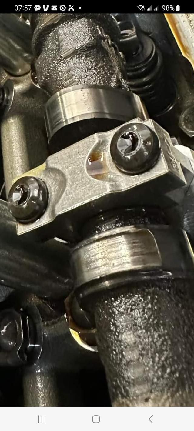 Close-up of a metal engine camshaft showing wear on the lobes and a bearing cap secured with two bolts.